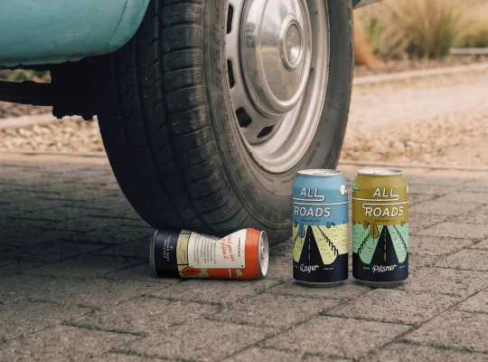 All Roads Brewing Co. brand photography showing cans on ground near vintage car wheel