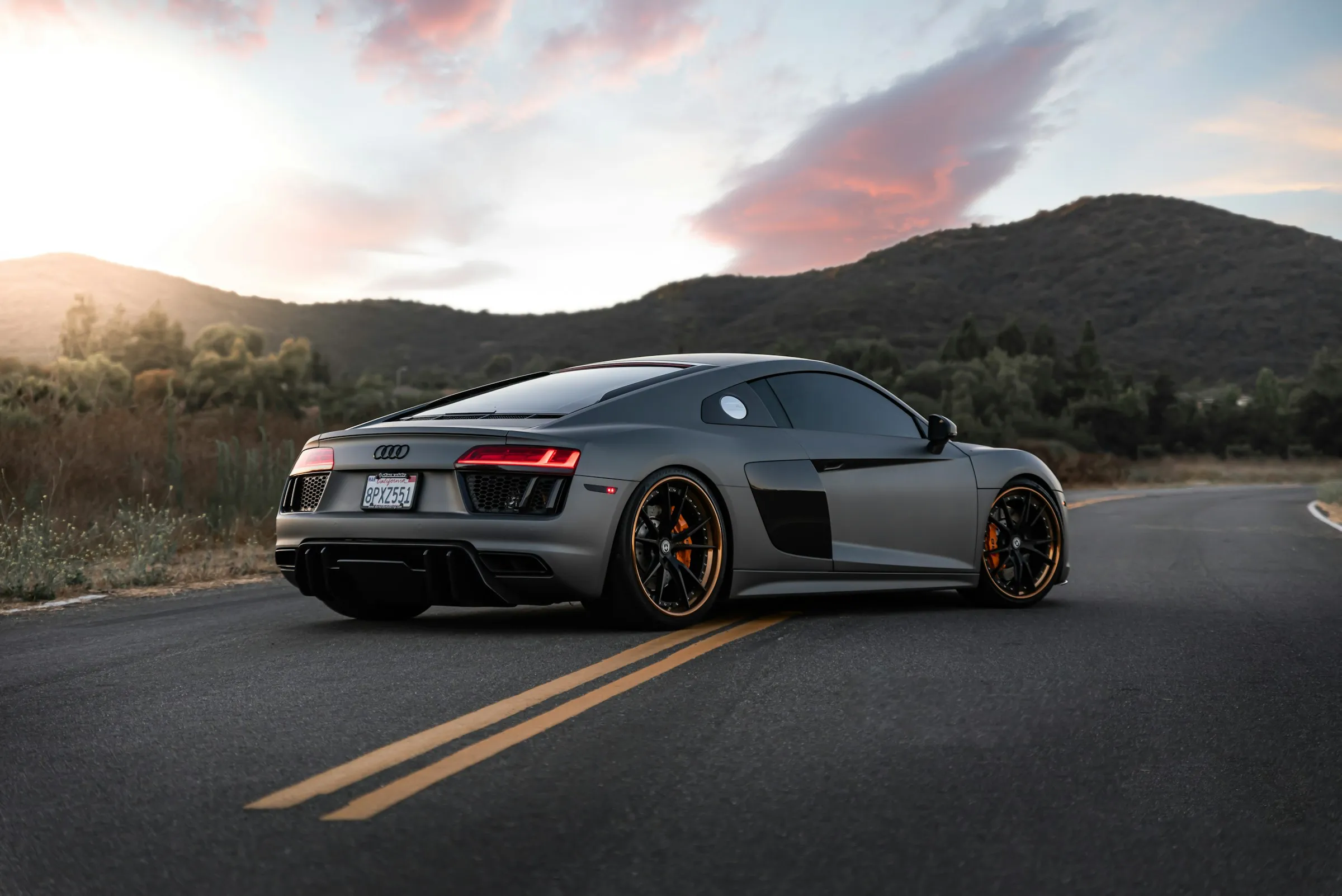 Gray Audi sports car parked on a mountain road at sunset, viewed from the rear with landscape in the background