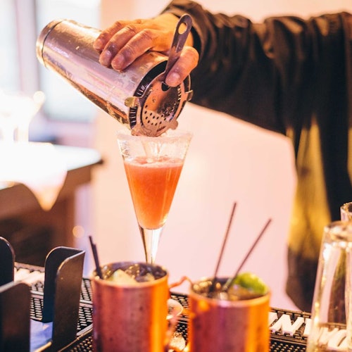 Person pouring a pink cocktail from a shaker into a glass, with two copper mugs and a bar in the background.