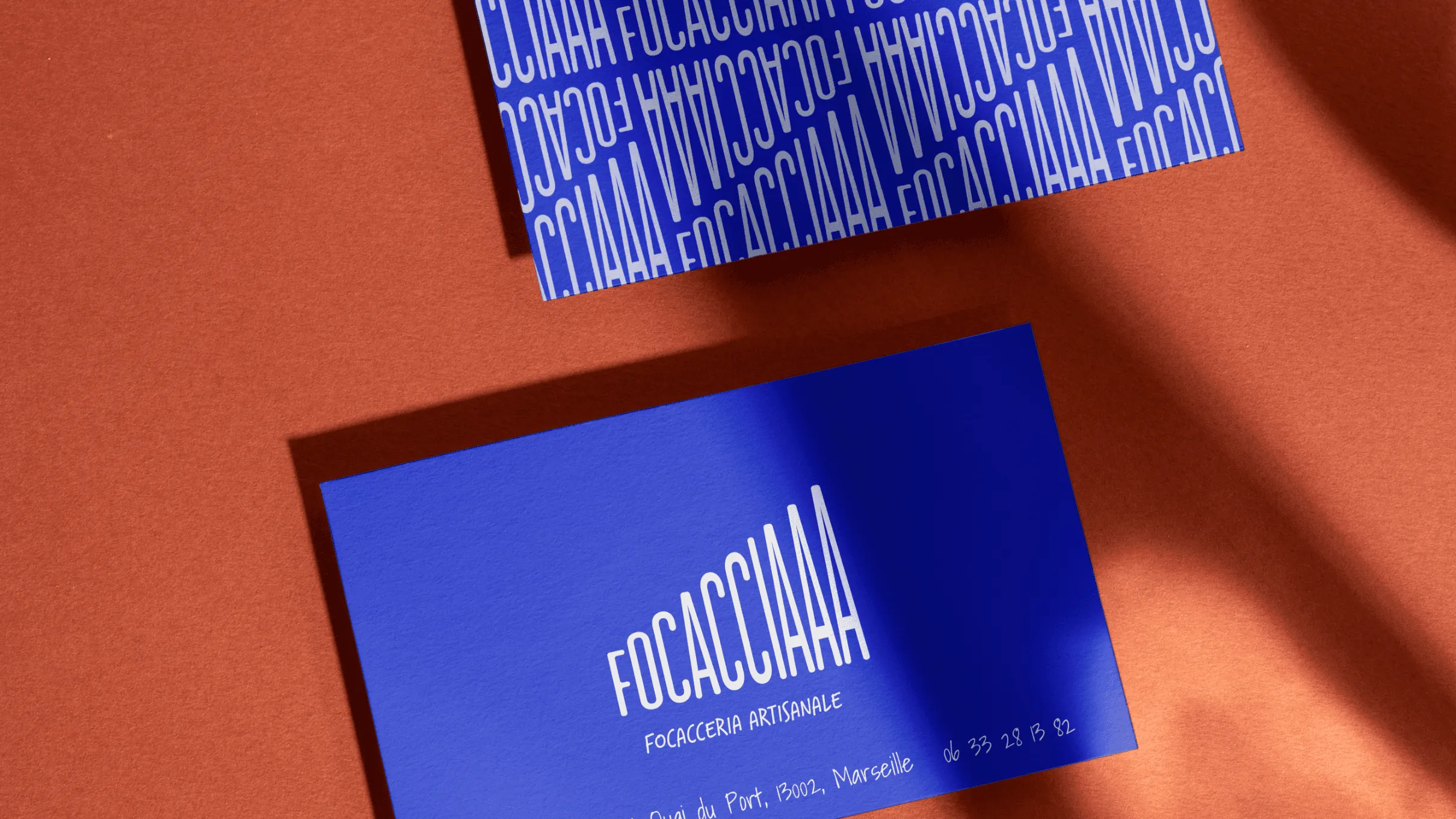 Deux cartes de visite bleues avec FOCACCIAAA, FOCACCERIA ARTISANALE et coordonnées, sur un fond texturé terracotta.