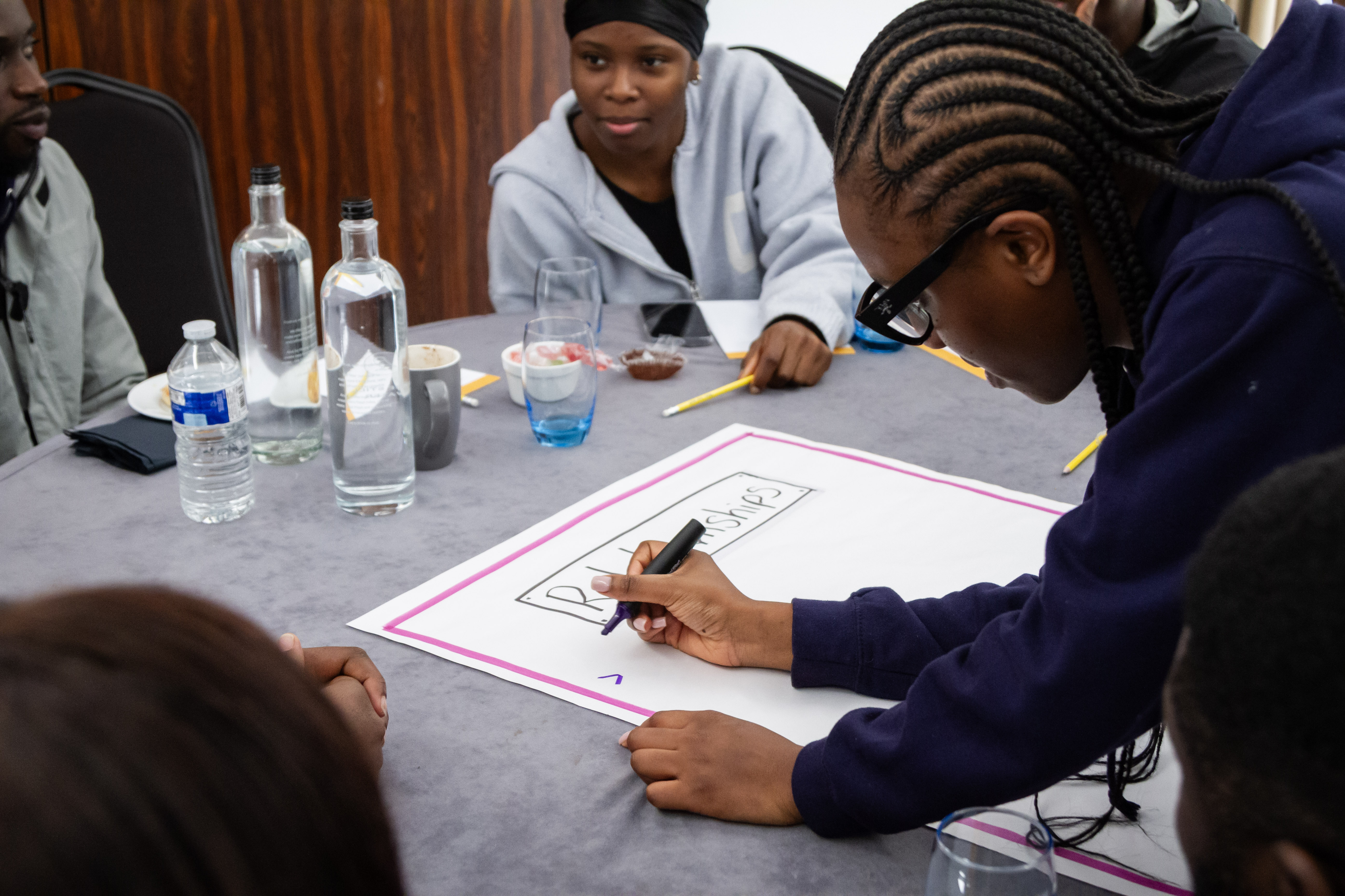 A person writing on a large sheet of paper at a table surrounded by others in a collaborative setting.