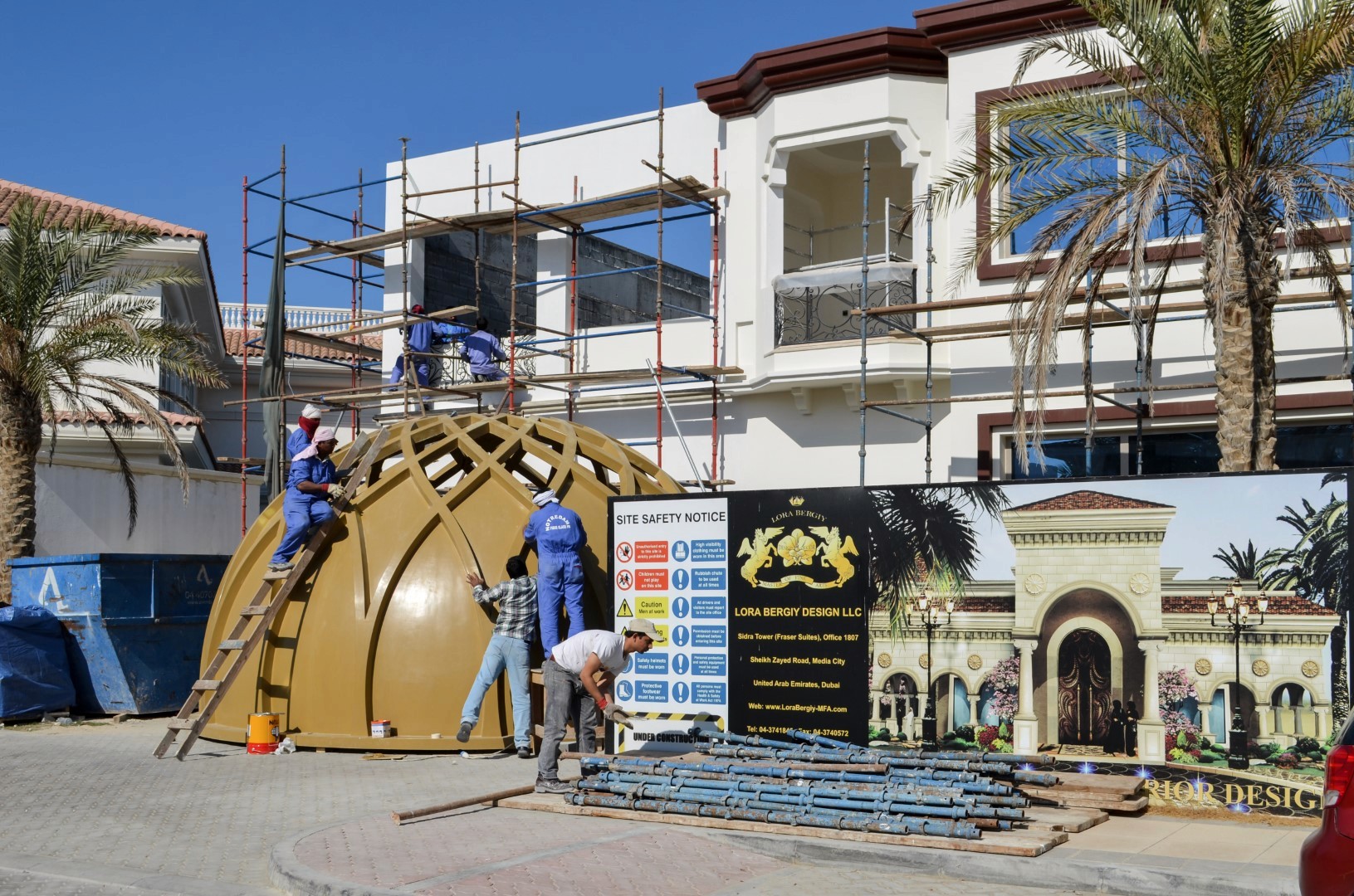 Construction workers in blue uniforms set up scaffolding beside a dome structure in front of a large, white residential building with palm trees, highlighting an ongoing construction project in a sunny location.