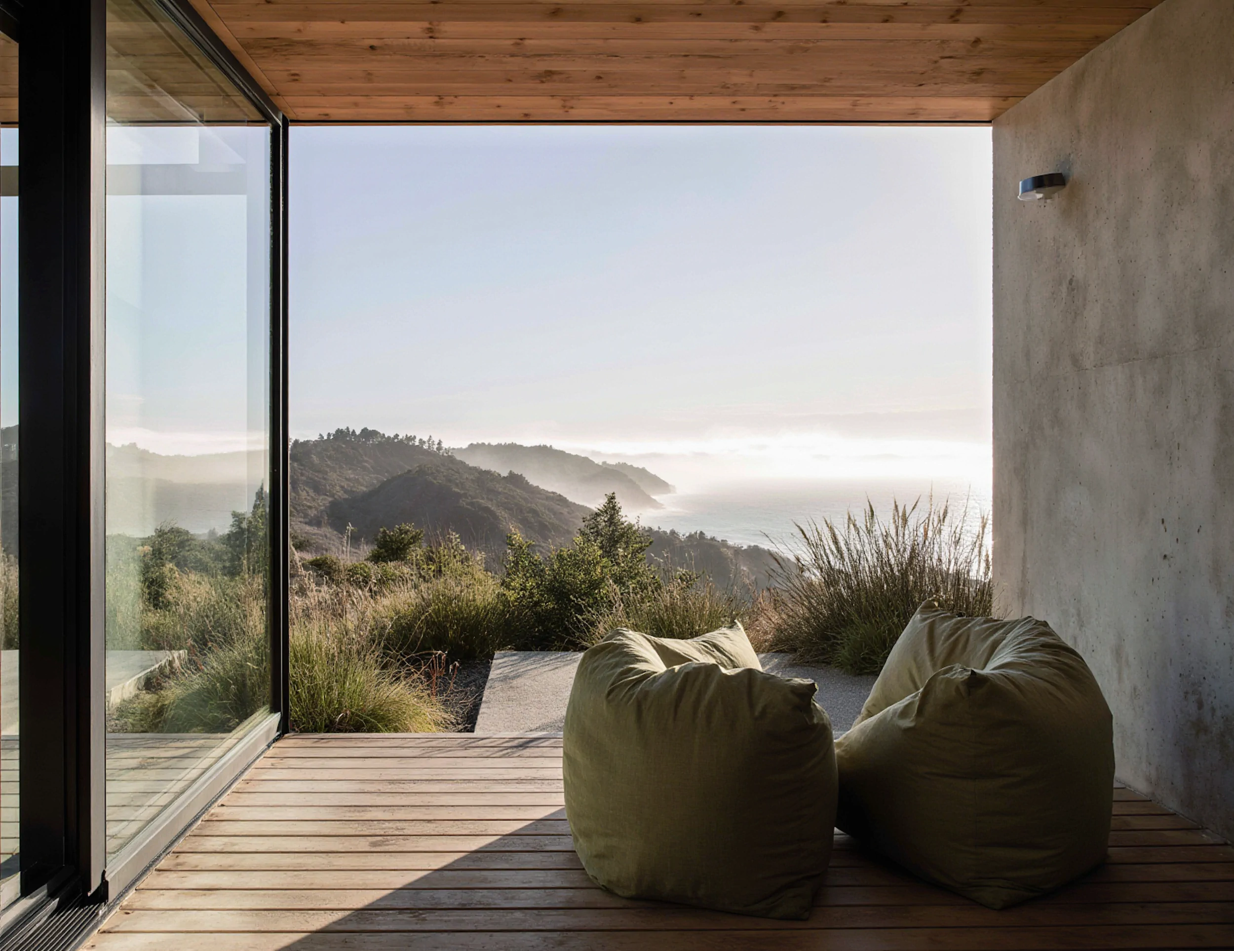 Modern concrete-and-wood room opening to a coastal overlook, with soft lounge seating facing expansive ocean views and morning fog.