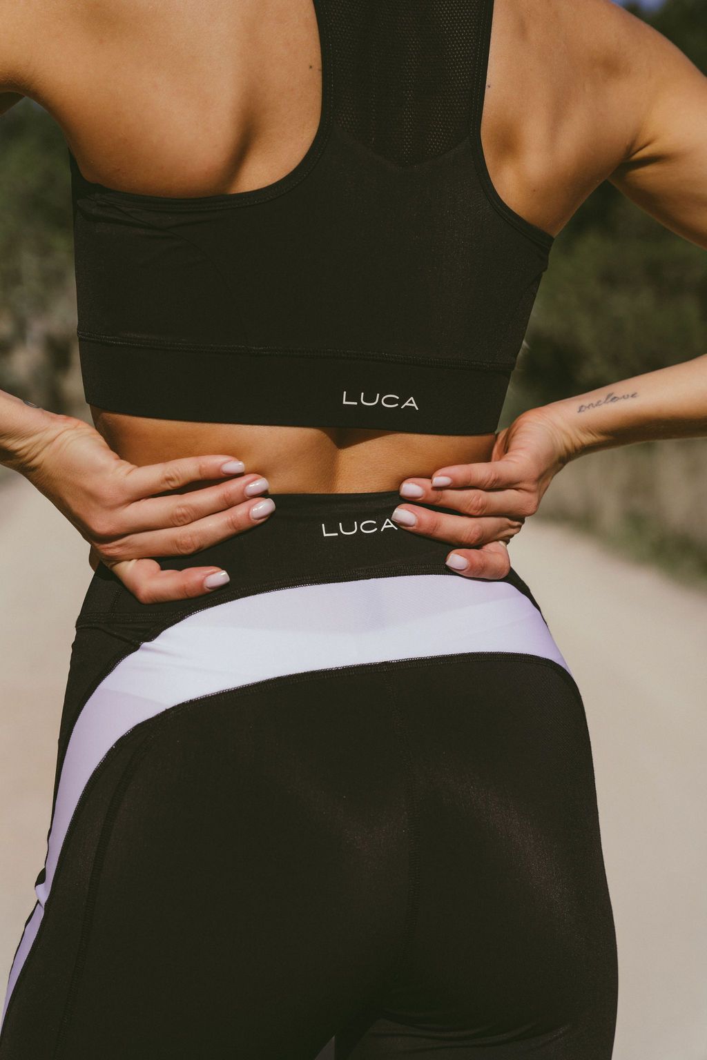 A close-up-view-of-a-woman-from-the-back-wearing-black-athletic-wear- that-shows-the-LUCA-brand-logo-on- the-cycling-bra-and-matching-high-waisted-leggings-with-a-thick-white- diagonal-stripe-The-woman's-hands- are-placed-on-her-lower-back-Ideal-for-cycling