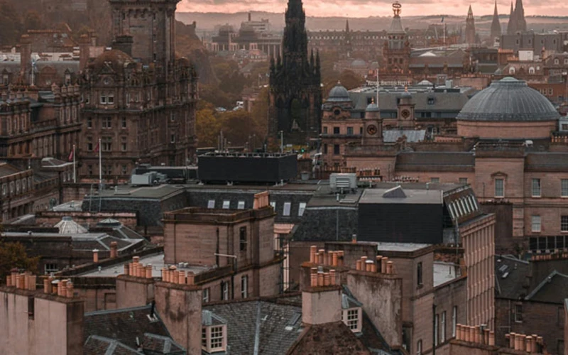 A panoramic view of Edinburgh’s cityscape at sunset, showing historic stone buildings, rooftops with chimneys, and prominent landmarks including the Scott Monument and the dome of the General Register House. The scene is bathed in warm, muted evening light.