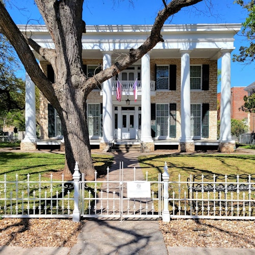 Una grande casa a due piani con alte colonne bianche, una recinzione in ferro e due bandiere americane all'ingresso.