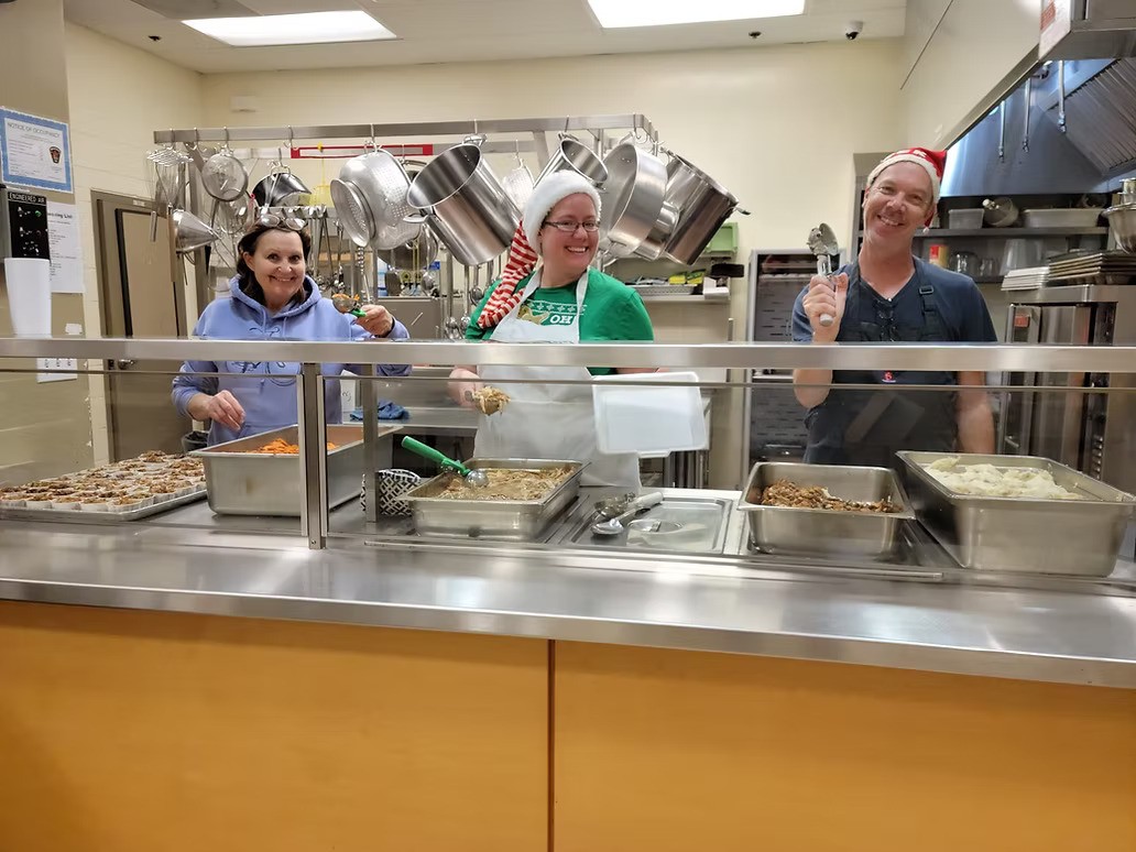 Three cheerful volunteers in festive Santa hats serve hot meals from a cafeteria-style kitchen, smiling behind a counter lined with trays of food including mashed potatoes, stuffing, sweet potatoes, and green beans. Stainless steel pots hang behind them, adding to the warm, communal holiday spirit.