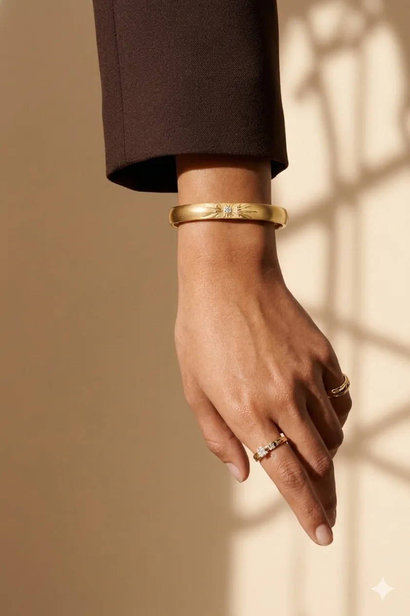 A hand adorned with elegant jewelry showcases a gold bracelet and two rings against a softly lit, neutral background.