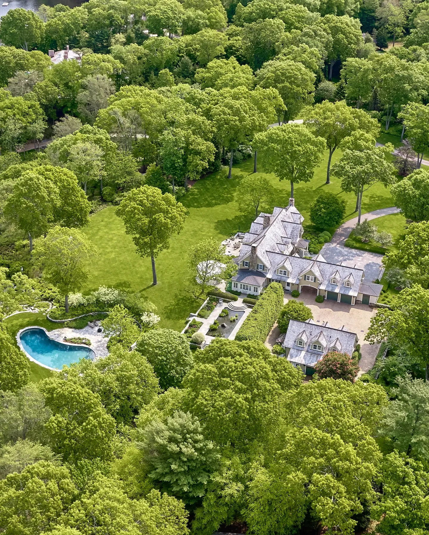 Aerial view of a sprawling estate with a large white house featuring steeply pitched roofs, multiple chimneys, and dormer windows, surrounded by lush green lawns, tall trees, and a circular driveway.