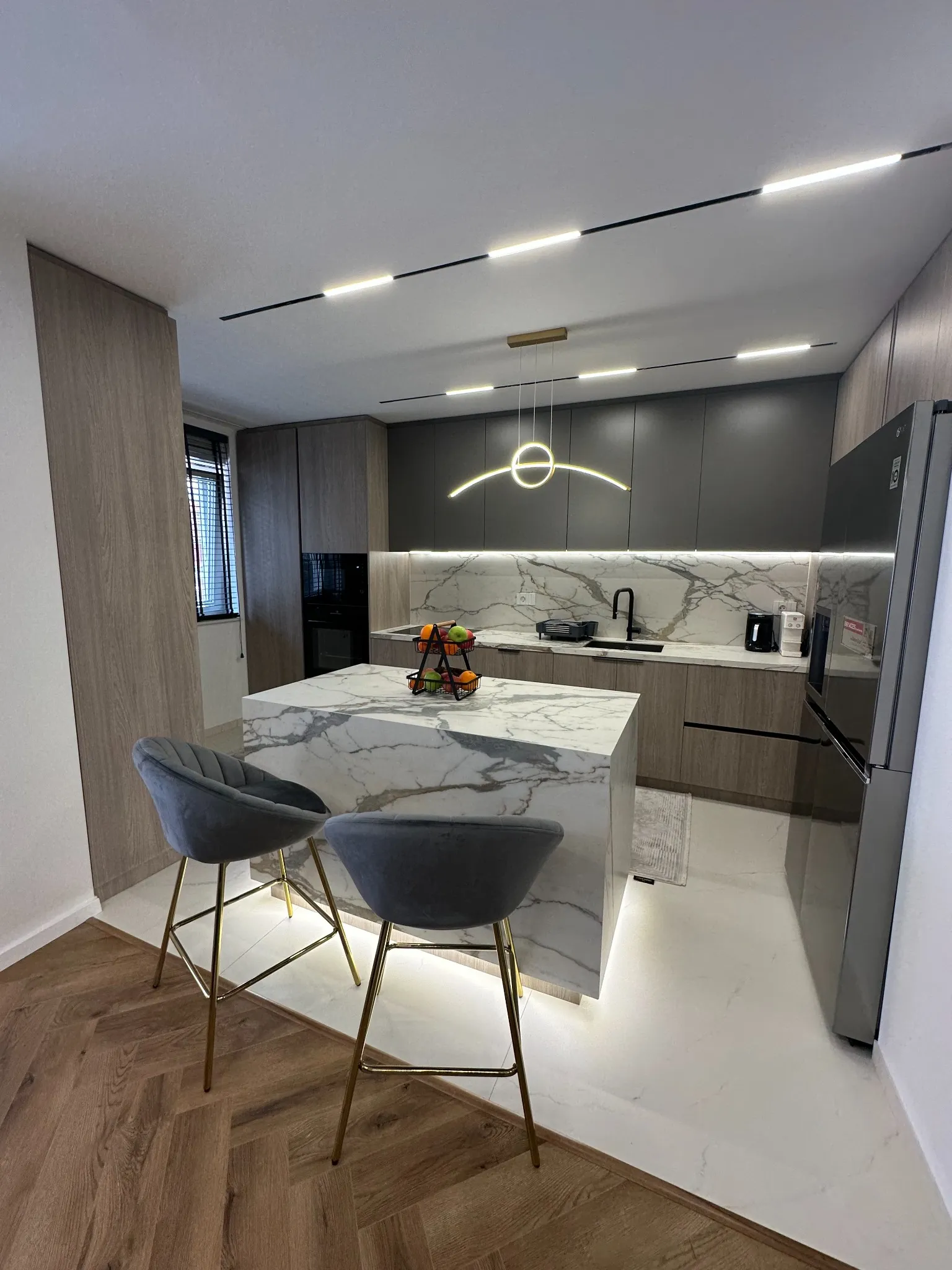 Contemporary kitchen in a Gostivar apartment designed by Terraplan Partners, showcasing a marble island, modern cabinetry, integrated lighting, and upholstered bar stools with gold accents.