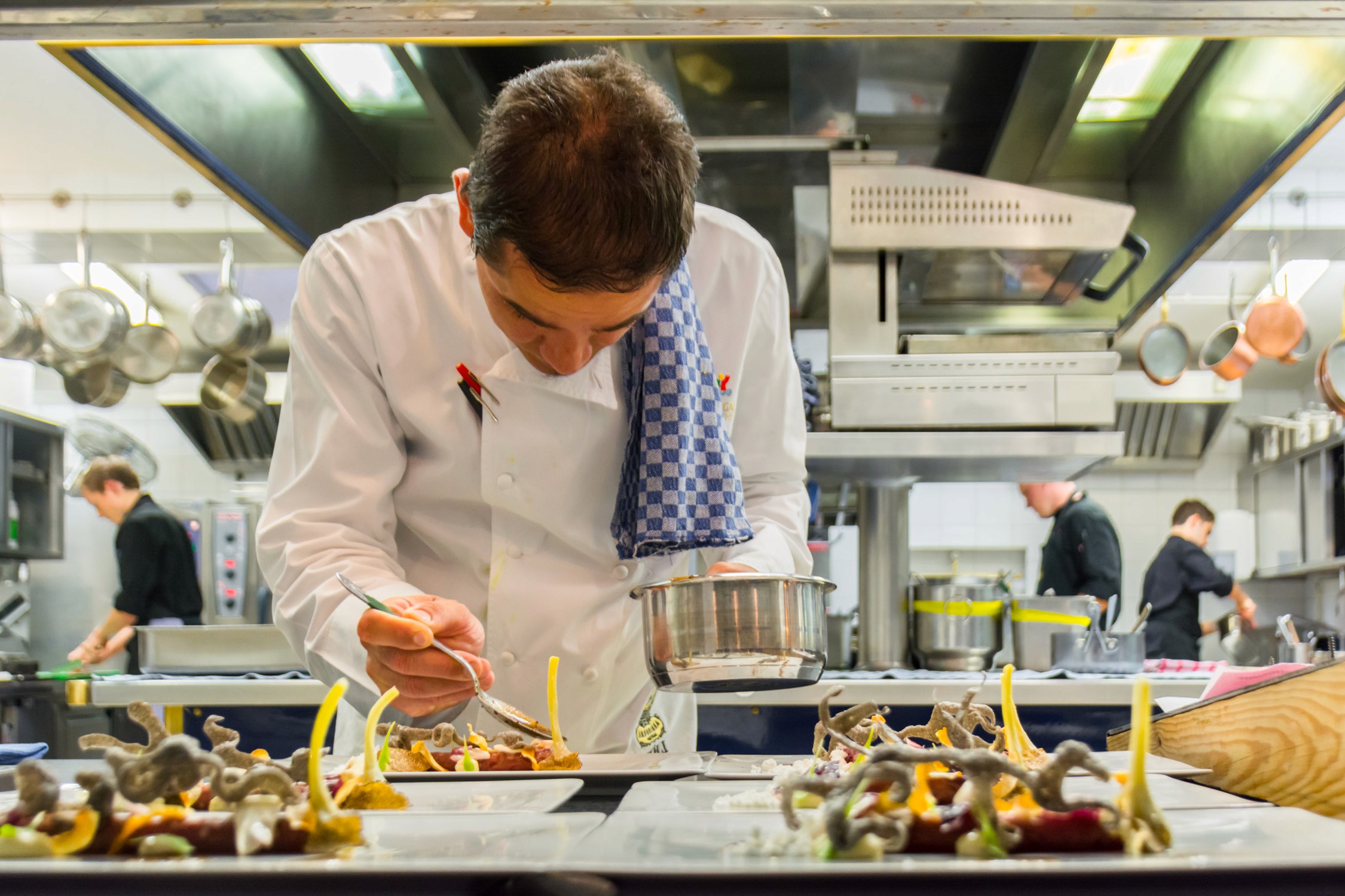 A chef in a white uniform and blue checkered towel prepares food at a stainless steel counter in a busy commercial kitchen.