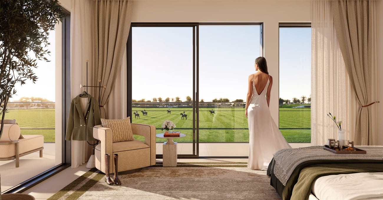 A woman wearing a white dress stands by a window, looking out at a field at the Grand Polo Club and Resort.