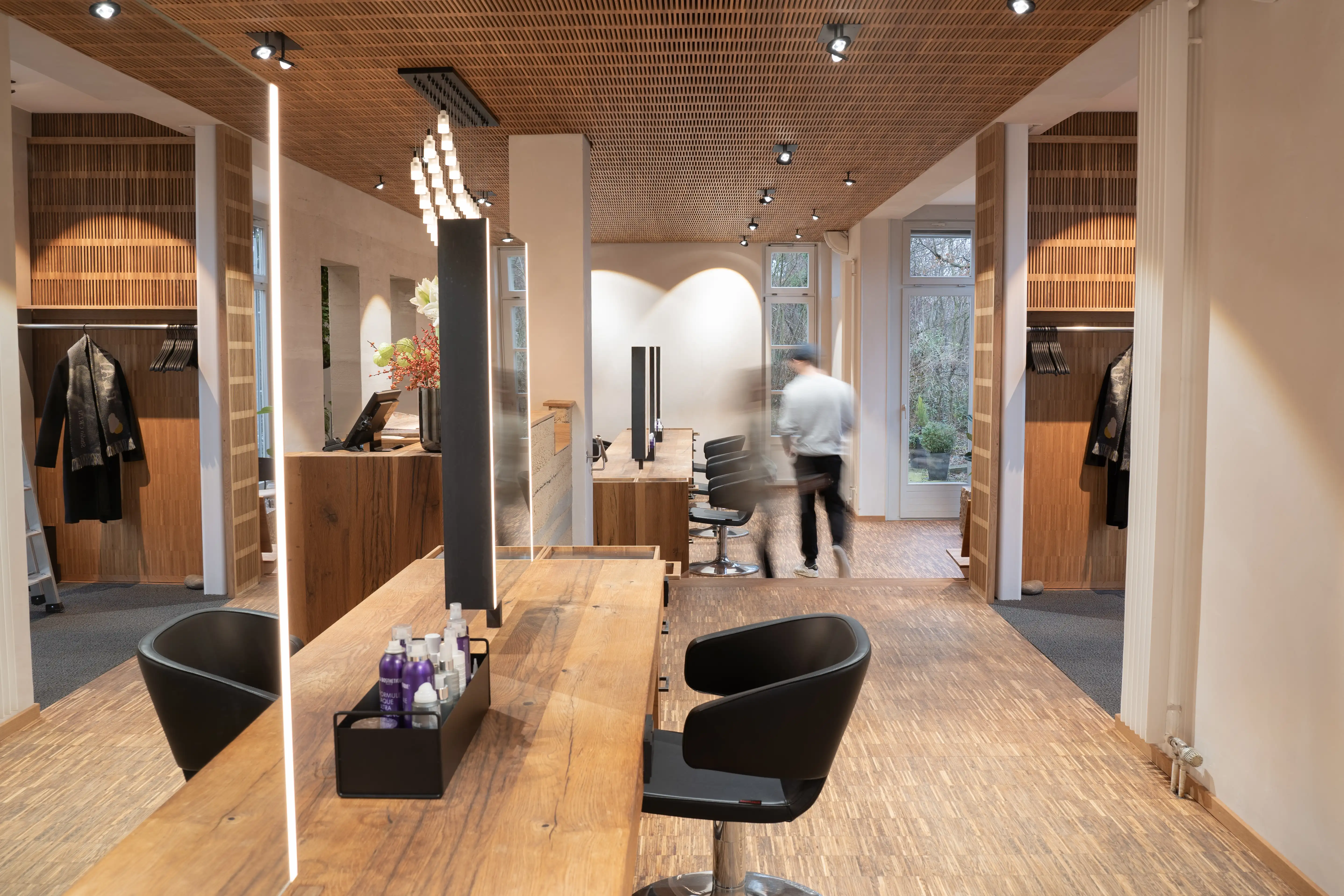 Overview of the EMPL FRISEURE salon with a long oak styling table, black chairs, illuminated mirror panels, wooden wardrobe niches and parquet flooring.