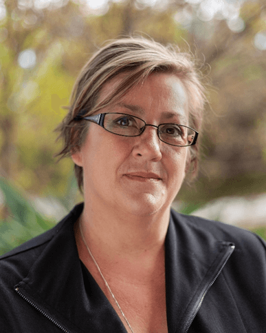A smiling man with glasses, curly hair, wearing a maroon hoodie, poses against a dark background.