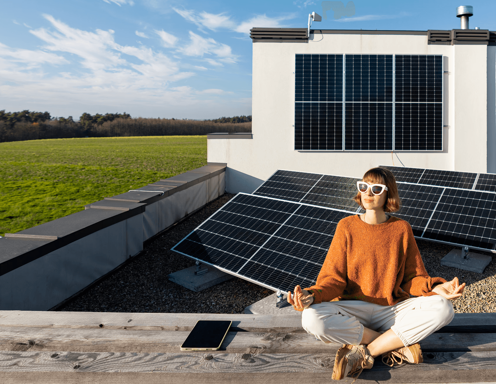 Eine Person sitzt mit gekreuzten Beinen in einer meditativen Haltung auf einem Dach, umgeben von mehreren großen Sonnenkollektoren, unter einem klaren blauen Himmel, wodurch erneuerbare Energien und nachhaltiges Leben hervorgehoben werden.