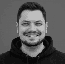 Woman in sunglasses poses against vibrant red background, wearing a beige blazer and black top. Sunlight casts dynamic shadows, creating a stylish, confident mood.