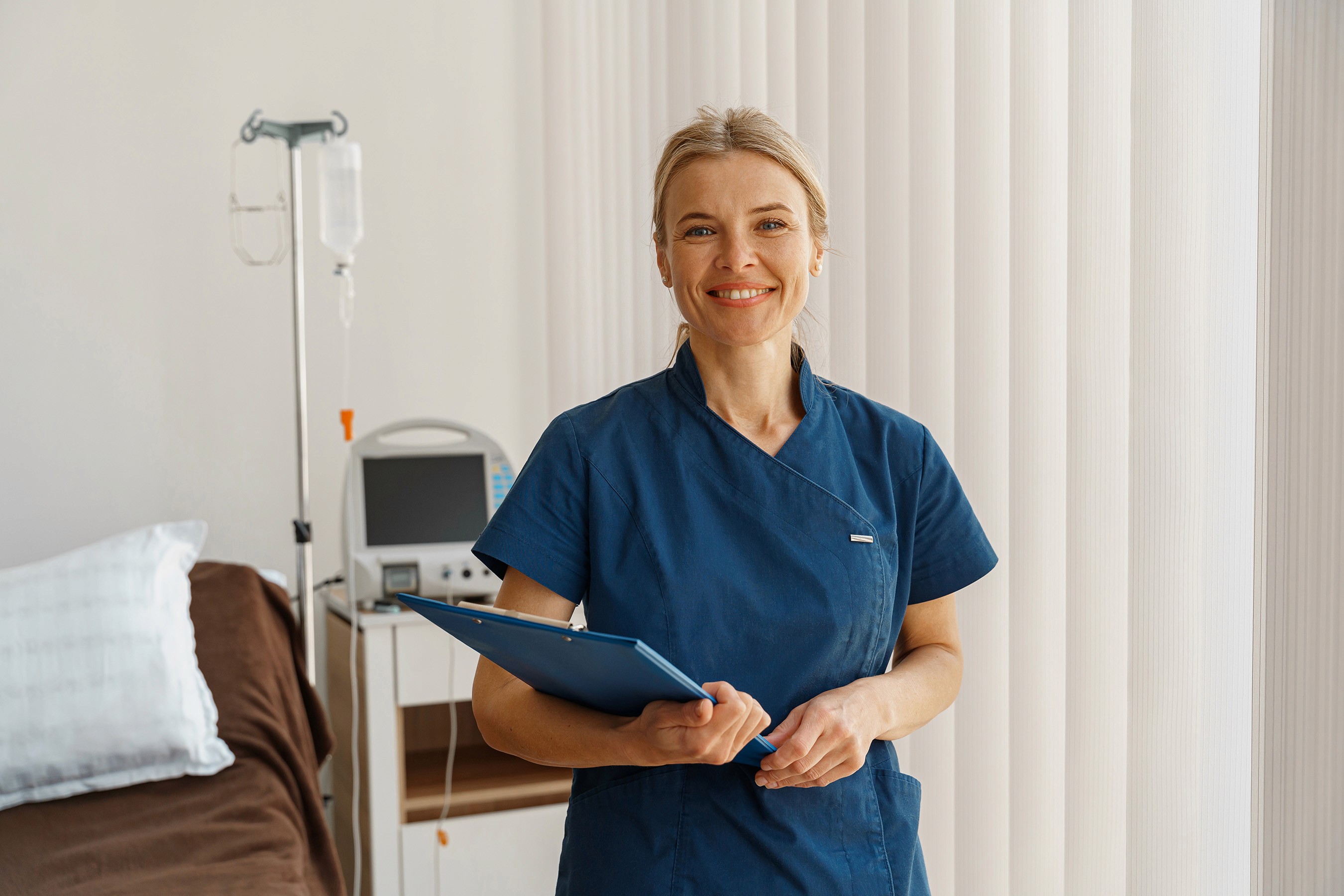 Smiling nurse in blue scrubs holding a clipboard in a medical room. A bed and medical equipment are in the background.