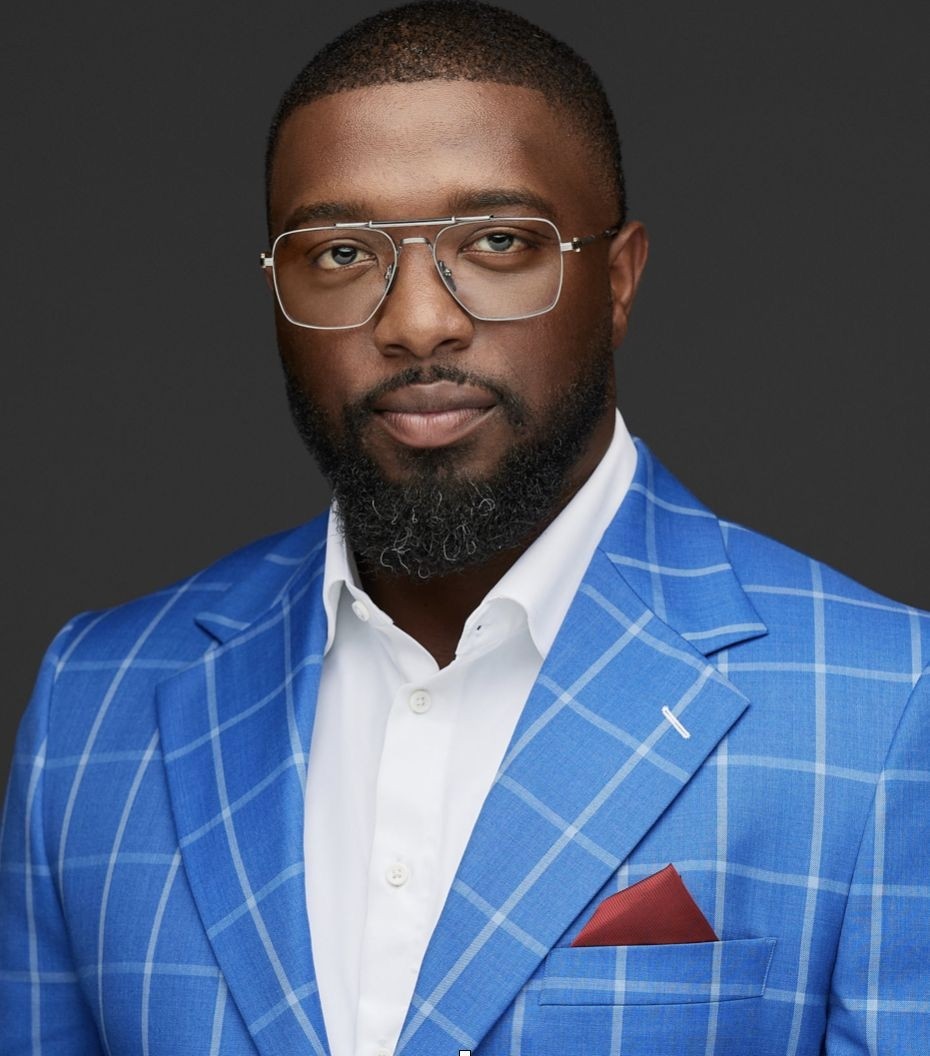 Portrait of a young man with wavy dark hair and a trimmed beard in a white sweater, soft light on a neutral background.