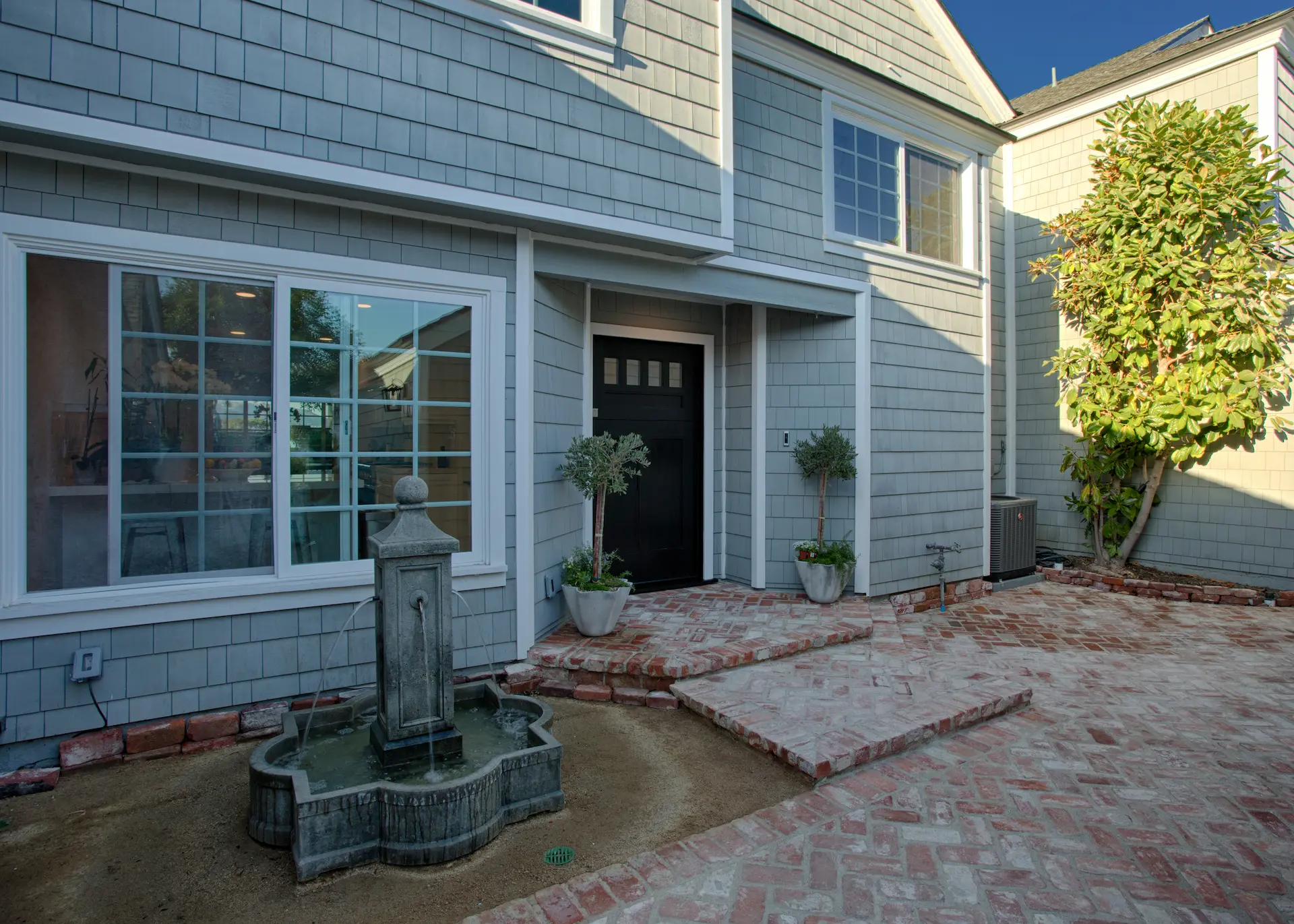 Brick courtyard with central fountain, front view of a Cape Cod style home, windows reflecting the sky. Photo by Chris Darnall