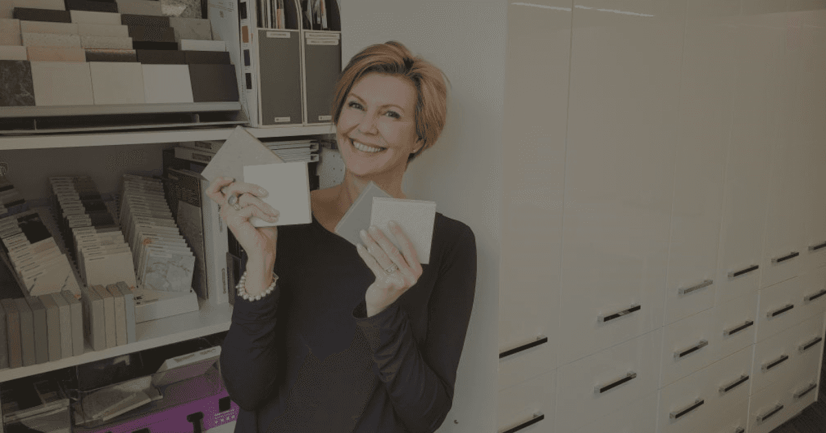 A fair skinned woman with blond hair holds square stone countertop samples in her hands in front of a large white cabinet that holds many other samples.