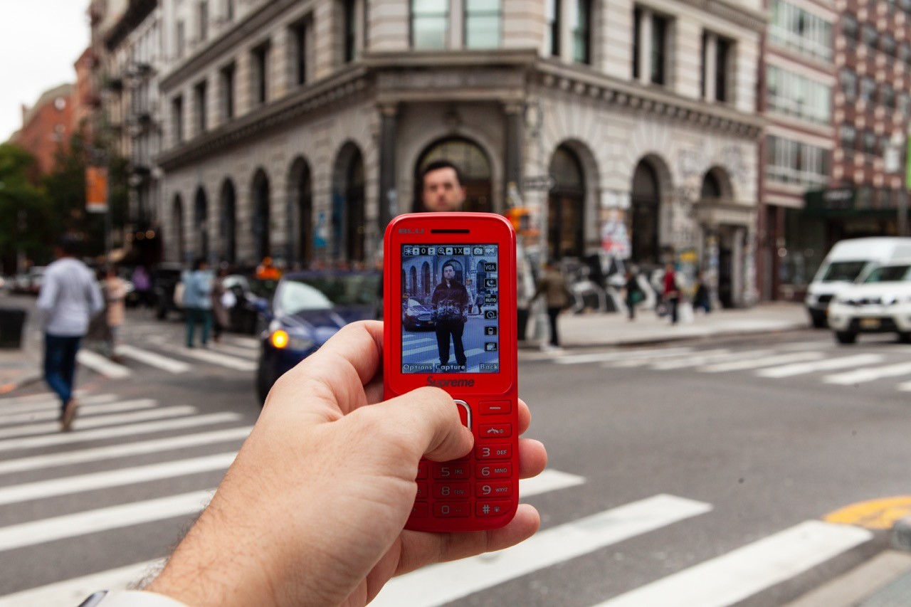 Hand holding a red feature phone showing a photo on screen, with a blurred city street in the background.