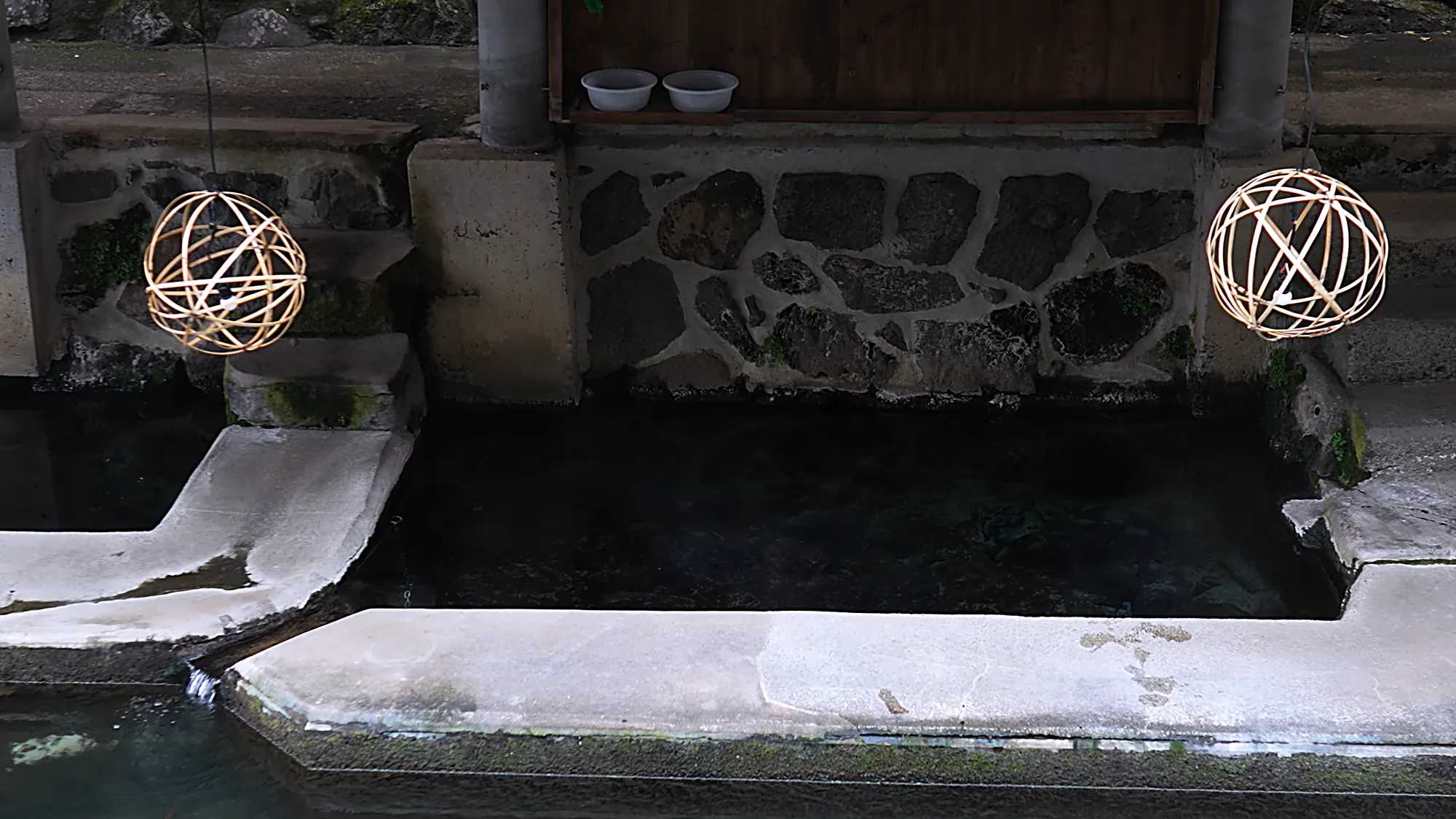 A riverside bathing platform at Kurokawa Onsen behind a stone wall, lantern spheres above