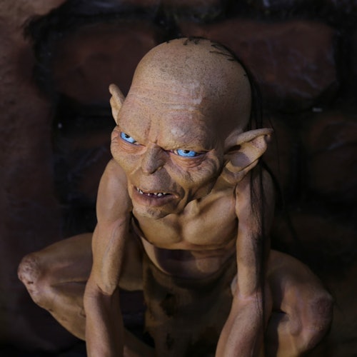 Eine Figur, die einem Kobold oder einer Kreatur gleicht, mit kahlem Kopf, spitzen Ohren, blauen Augen und scharfen Zähnen, die in einem schwach beleuchteten steinernen Umfeld hockt.