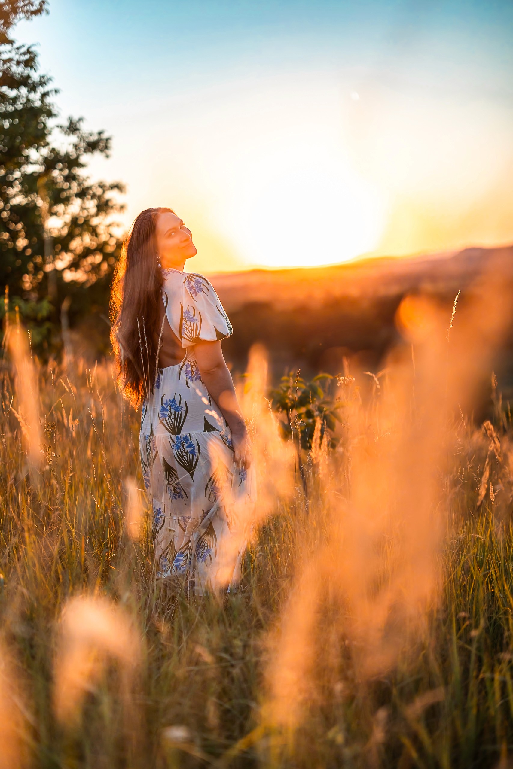 Hosszú hajú nő fehér virágmintás ruhában feltekint az égre aranyóra naplementében, búzamező és fák között Szécsényben, természetes arany fényben