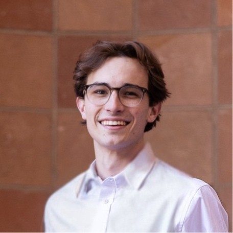 A young man with glasses smiles in front of a wooden wall, wearing a light-colored shirt.