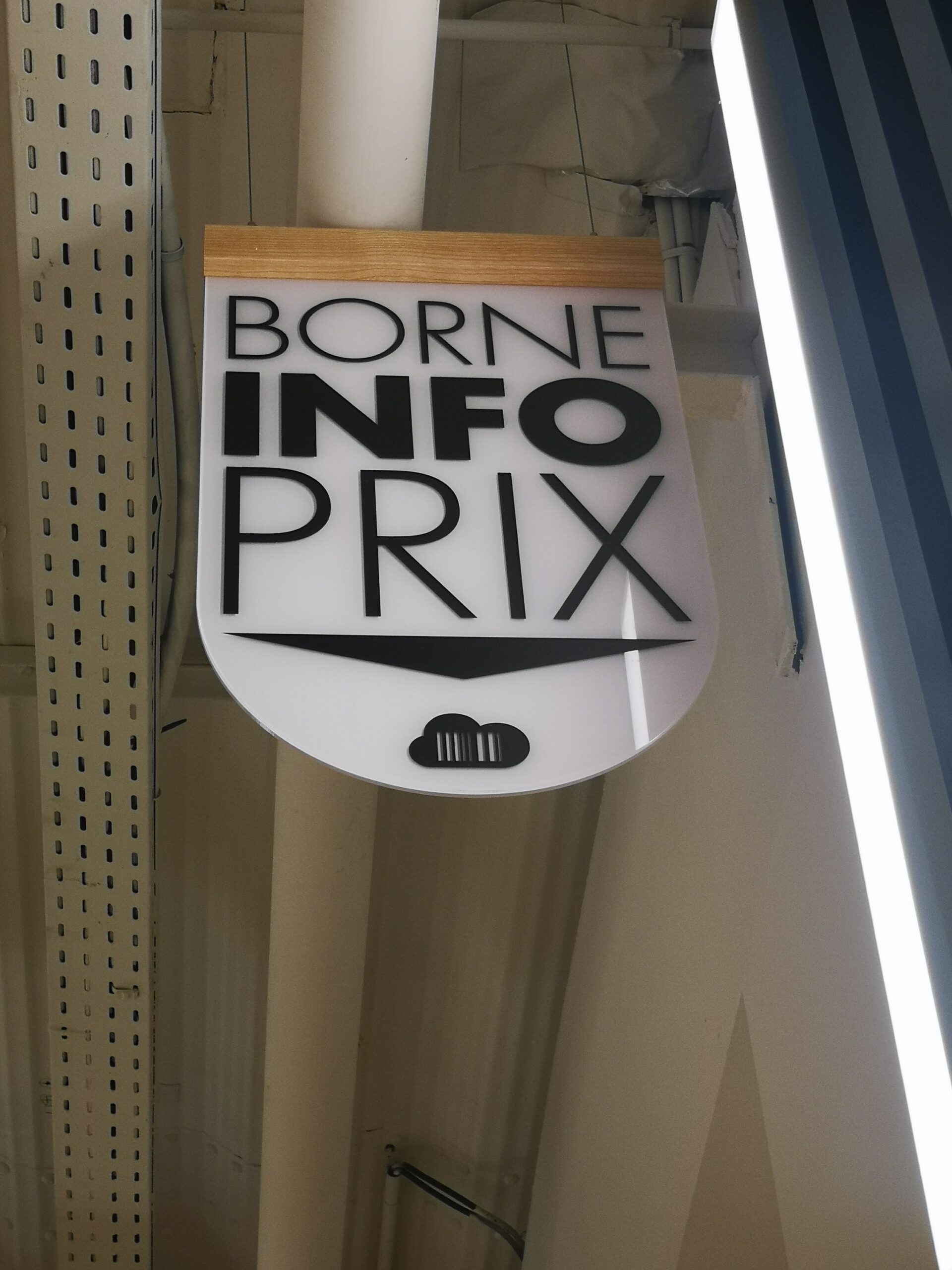 Signalétique Interne suspendue avec texte noir indiquant « Borne Info Prix » sur fond blanc, avec une petite icône de code-barres en dessous. Structure industrielle du plafond visible au-dessus.