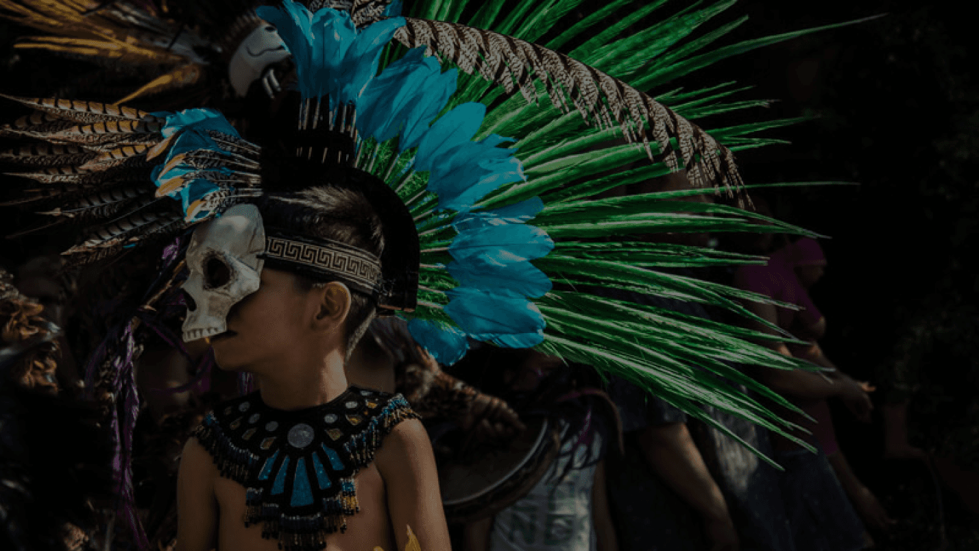 Fotografía de un niño con indumentaria tradicional de danzante mexica, portando un copilli o tocado de plumas y una máscara ceremonial.
