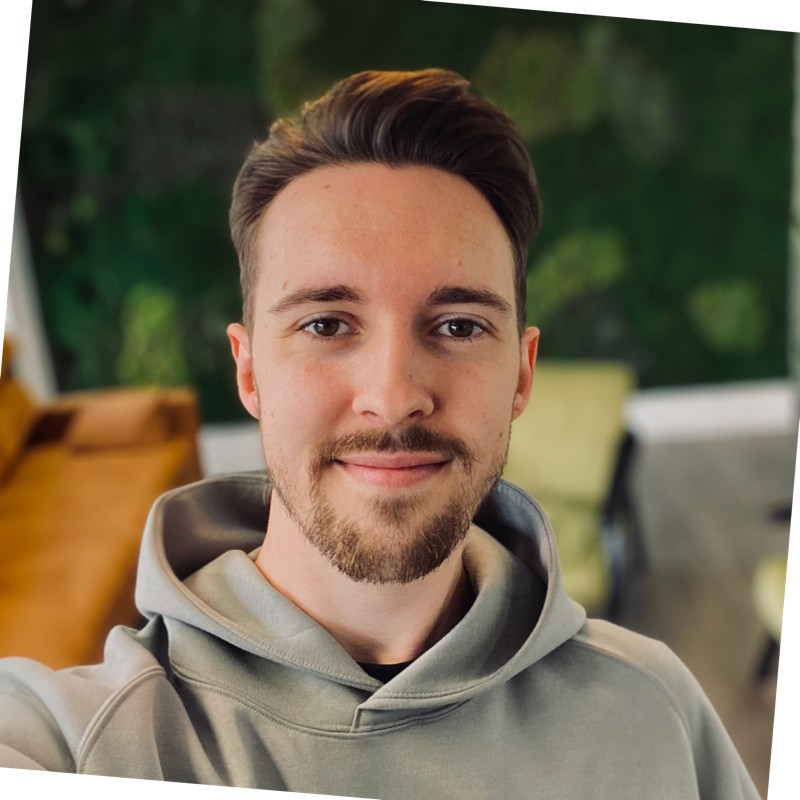 Close-up portrait of a person wearing a light grey hoodie, facing the camera indoors. The background is softly blurred and includes green wall décor, a tan sofa, and seating, creating a modern office or lounge setting with natural-looking light.