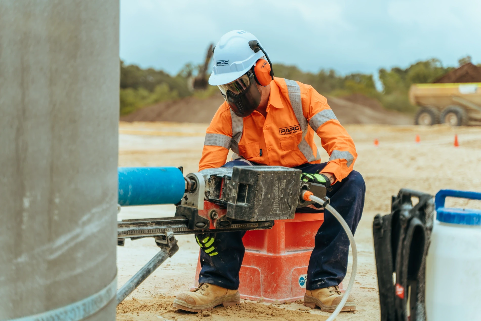 Worker in orange high-visibility clothing, ear muffs and a respirator operating a core drill against a concrete pile