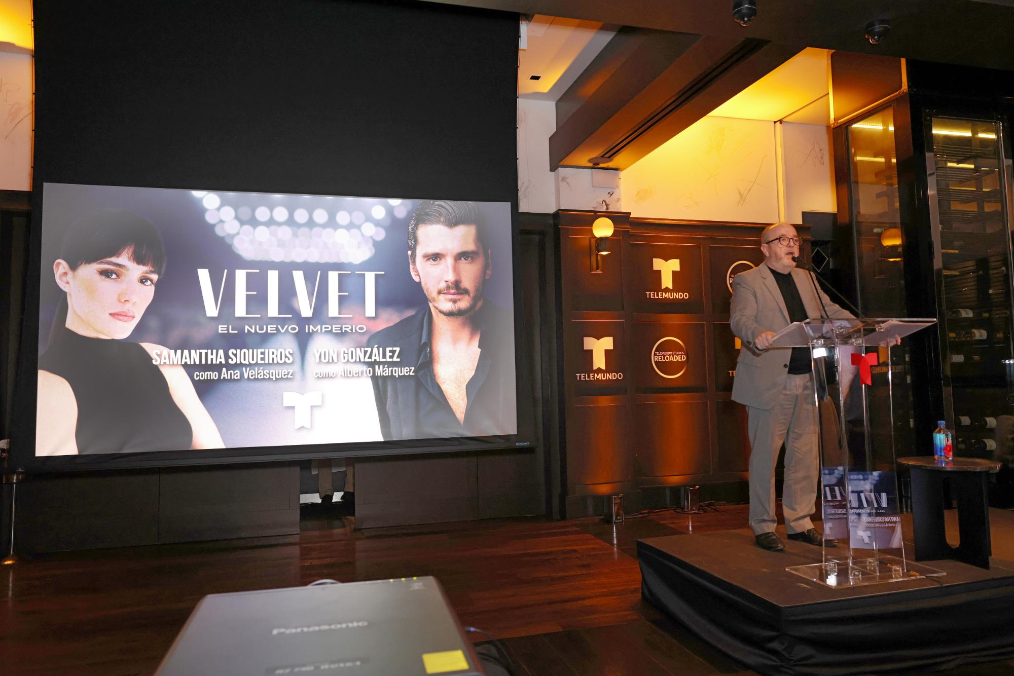 A speaker presents beside a large screen showing show artwork in an intimate venue setting.