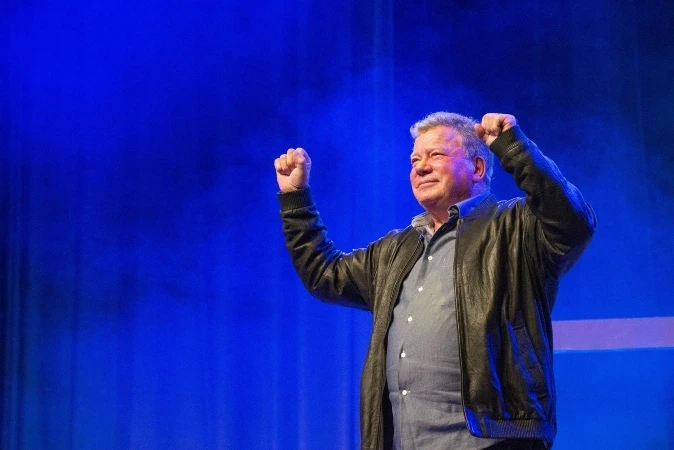 An older William Shatner in a leather jacket stands on stage with his arms raised in celebration against a blue-lit background.