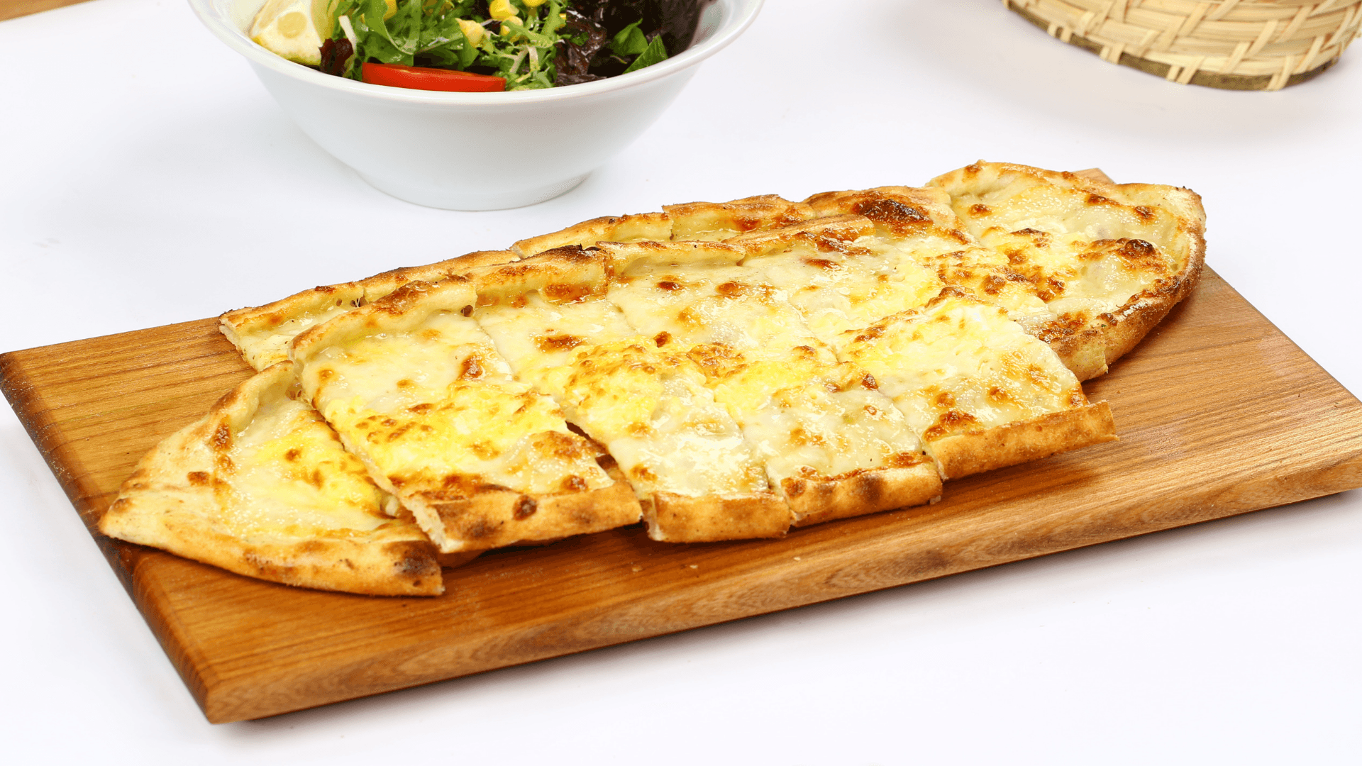 A freshly baked Turkish cheese pide (flatbread) served on a wooden board, with melted cheese topping and a side salad in the background.