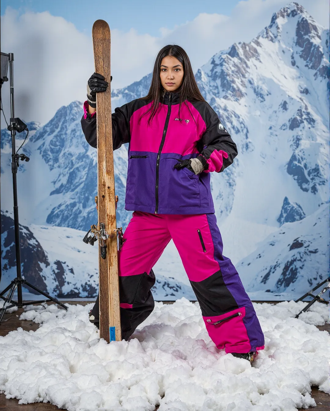 Vibrant pink and purple retro ski outfit with wooden ski against snow-capped mountain backdrop in a professional studio setting