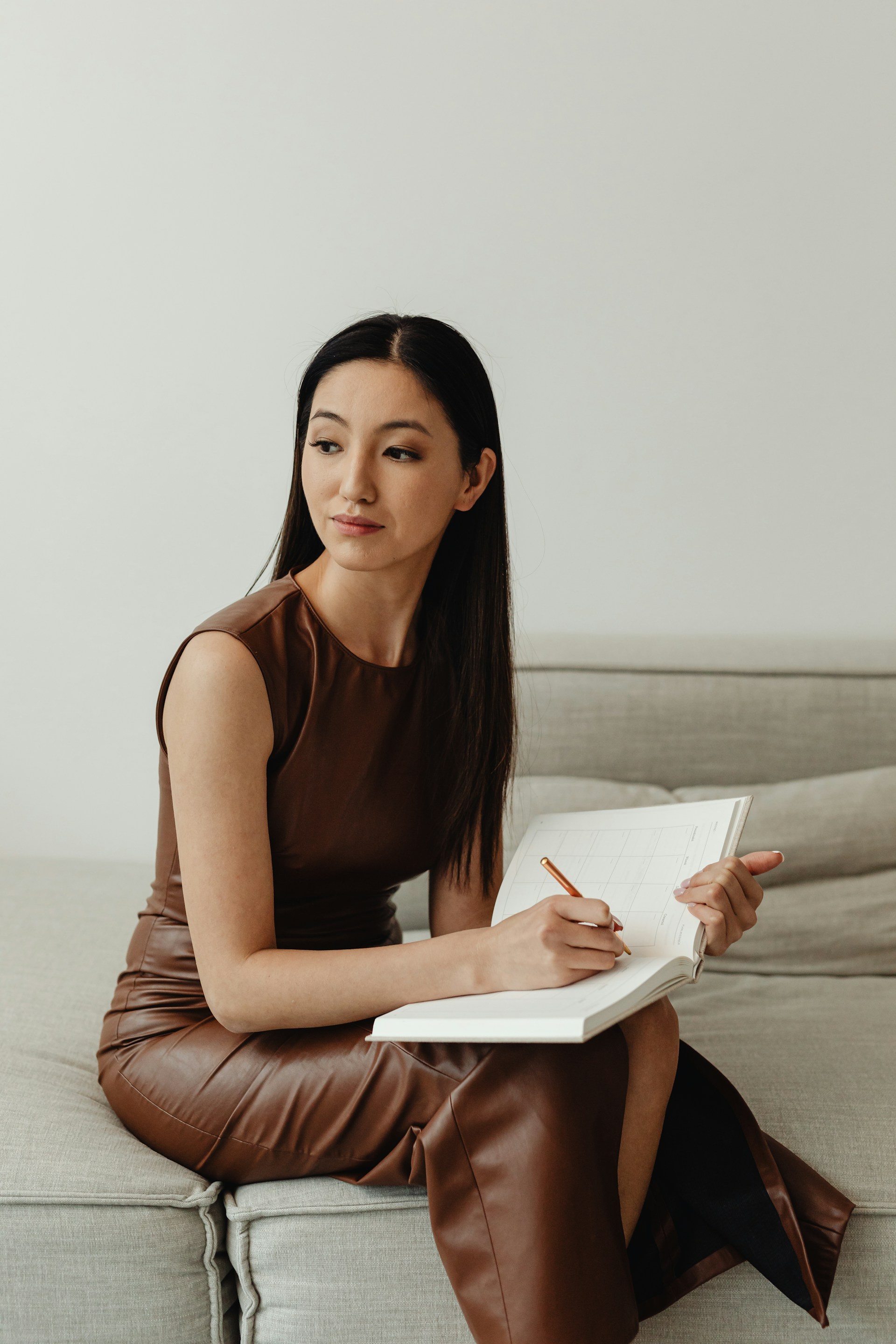 Woman sits on a sofa, writing in a planner and looking thoughtfully to the side.