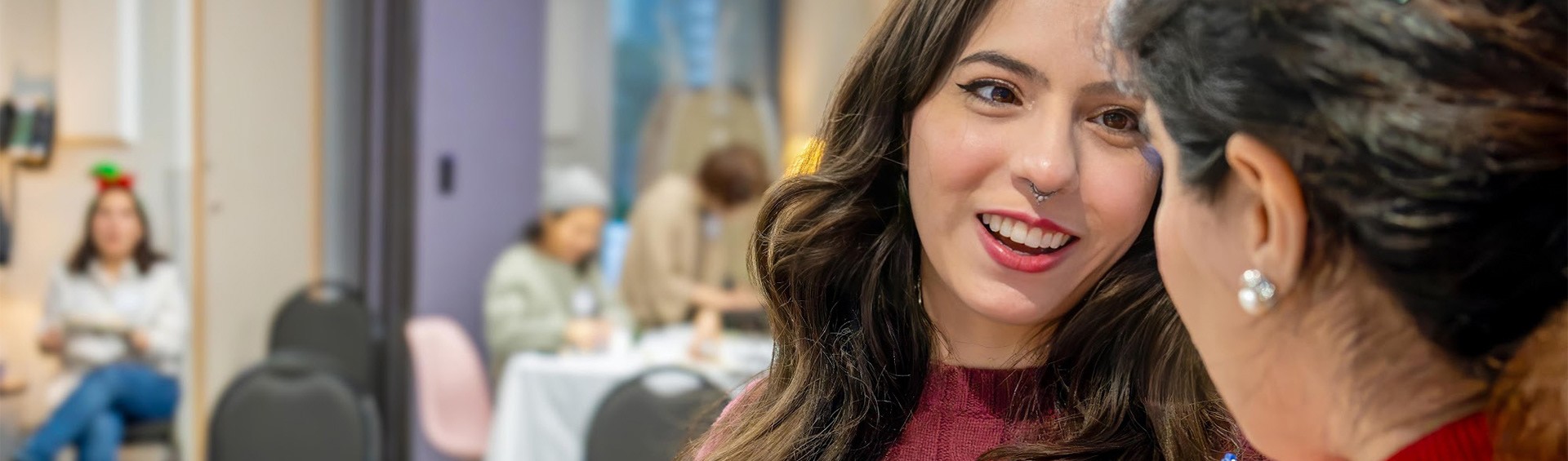 Close-up of two professional women in conversation in a contemporary office space. The woman facing the camera has long wavy brown hair, wears a burgundy knit top and nose ring, and displays an engaged, friendly expression. Another woman with short dark hair and pearl earrings is shown from behind. The bright, modern workspace features other colleagues working at desks in the background.