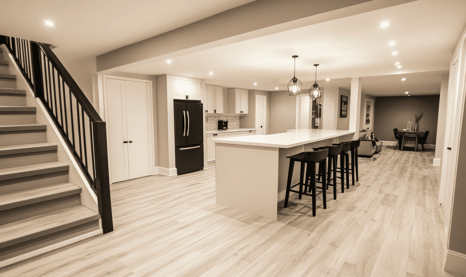 A spacious open-concept basement with a large white kitchen island featuring black bar stools. The area includes wood-look flooring, a black refrigerator, and modern pendant lighting, with stairs leading to another level in the background.