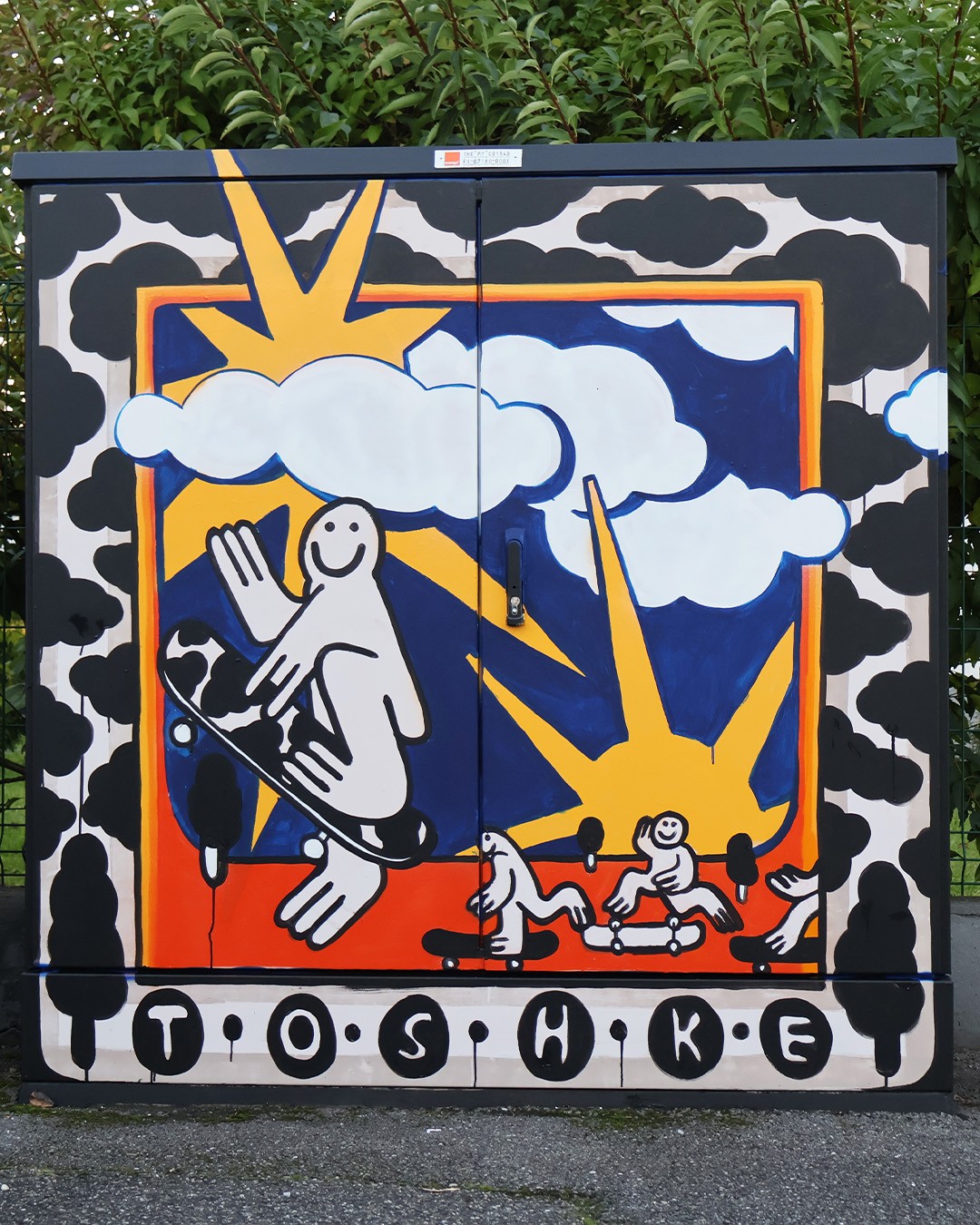 A brightly painted electrical box featuring joyful black-and-white cartoon skaters on an orange floor beneath yellow suns and clouds, framed by clouds and trees, with “TOSHKE” signed at the bottom.