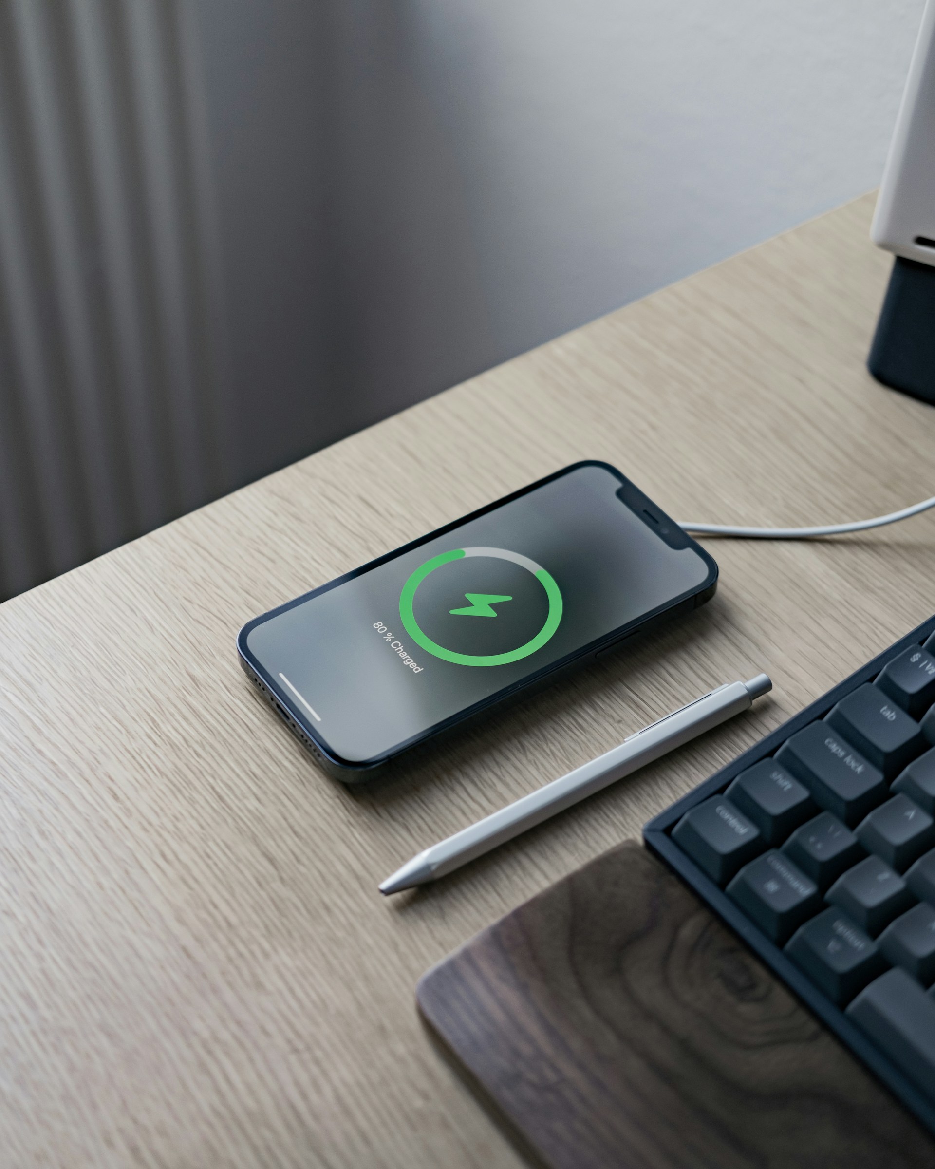 A smartphone with a glowing green **wireless charging** circle is resting on a light wooden desk next to a silver stylus pen and a dark keyboard.