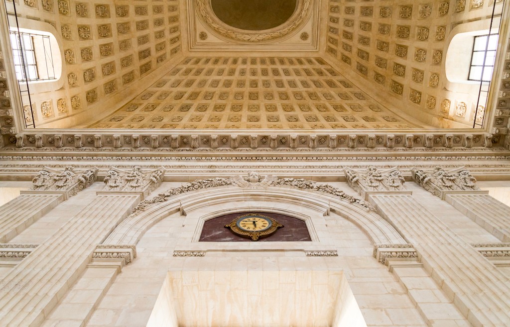 A person wearing headphones sits at a desk with a large curved monitor displaying graphic design software, surrounded by a laptop, smartphone, and potted plant, creating a modern workspace setup.