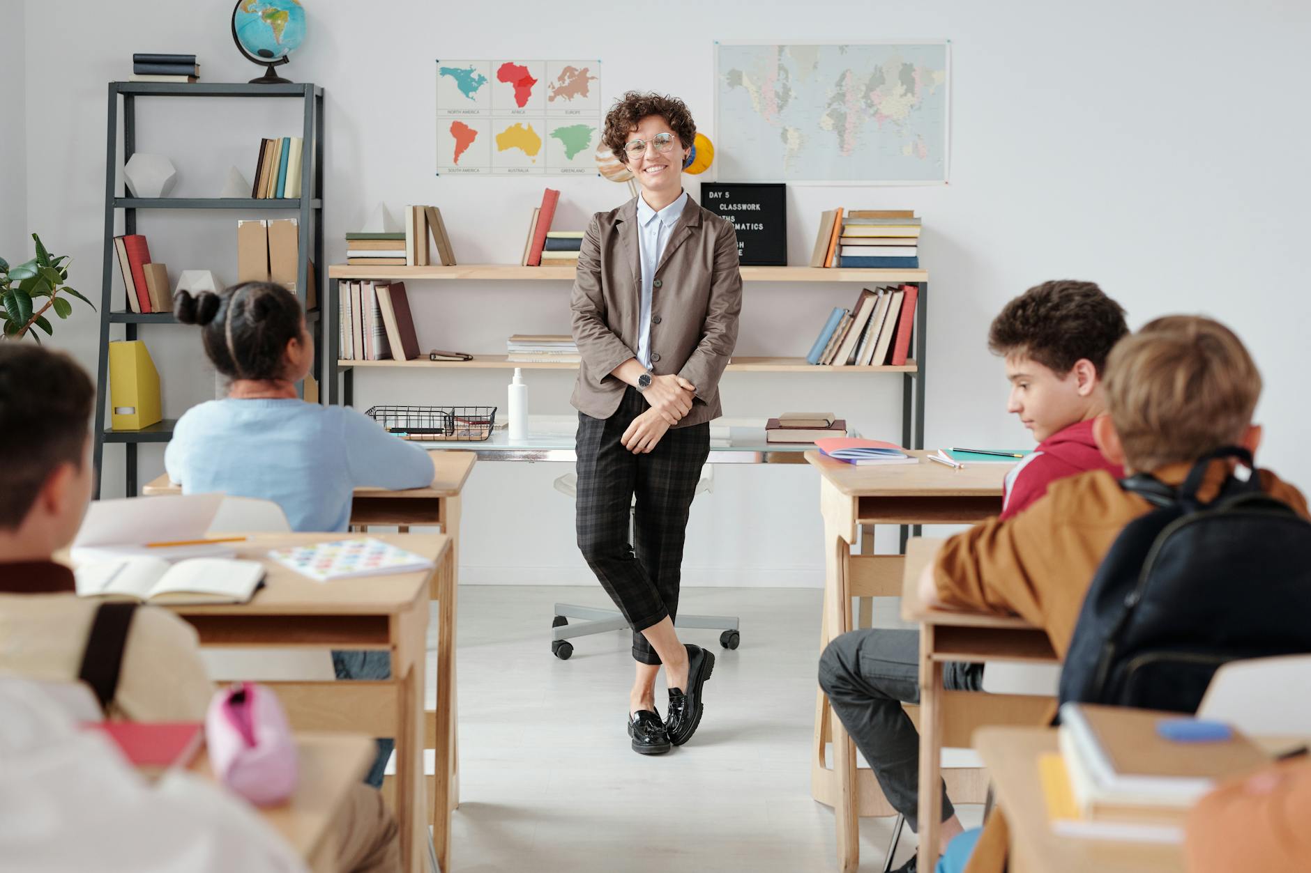 A teacher stands at a whiteboard calmly gesturing toward a set of classroom rules while students listen attentively.