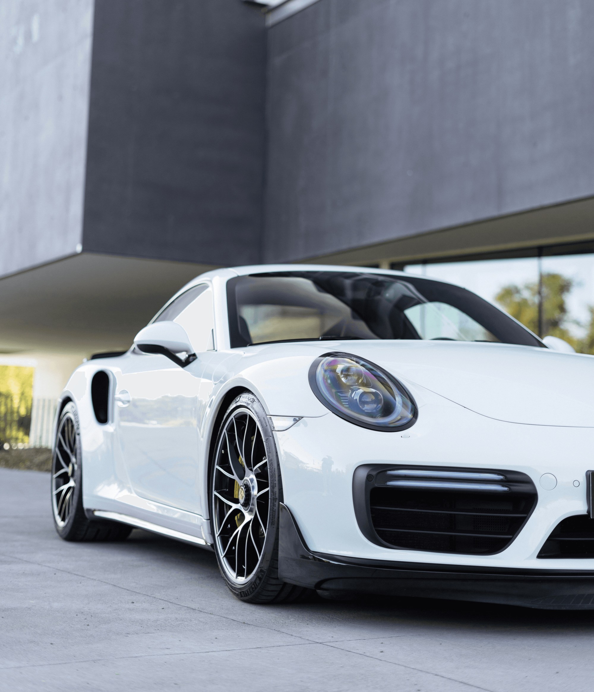 A sleek, white sports car with black alloy wheels is parked in front of a modern, minimalist concrete building, reflecting a sunny day.