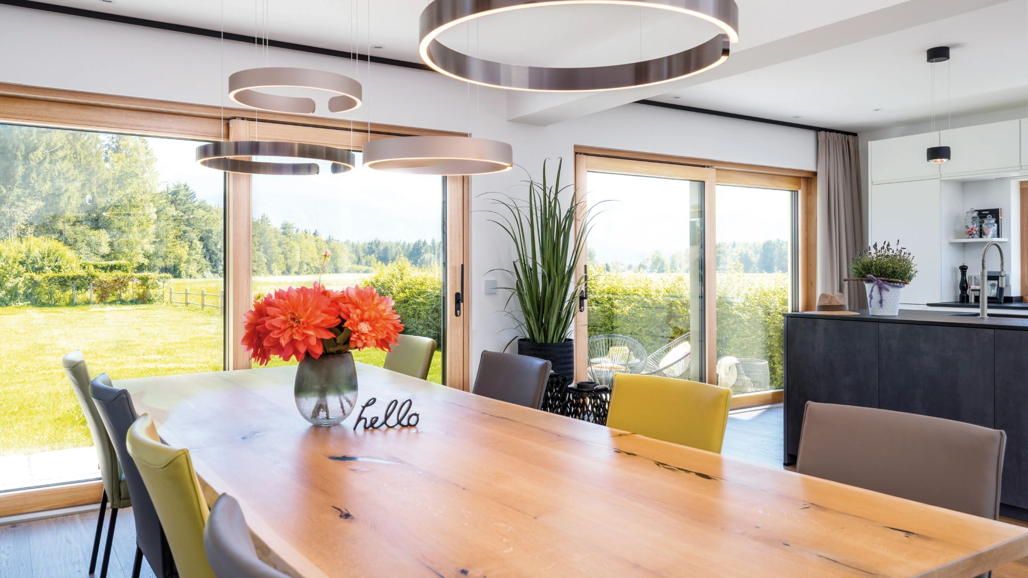 Modern Esszimmer mit Holztisch, bunten Stühlen, floraler Dekoration und ringförmigen Hängeleuchten vor Panoramafen stern.