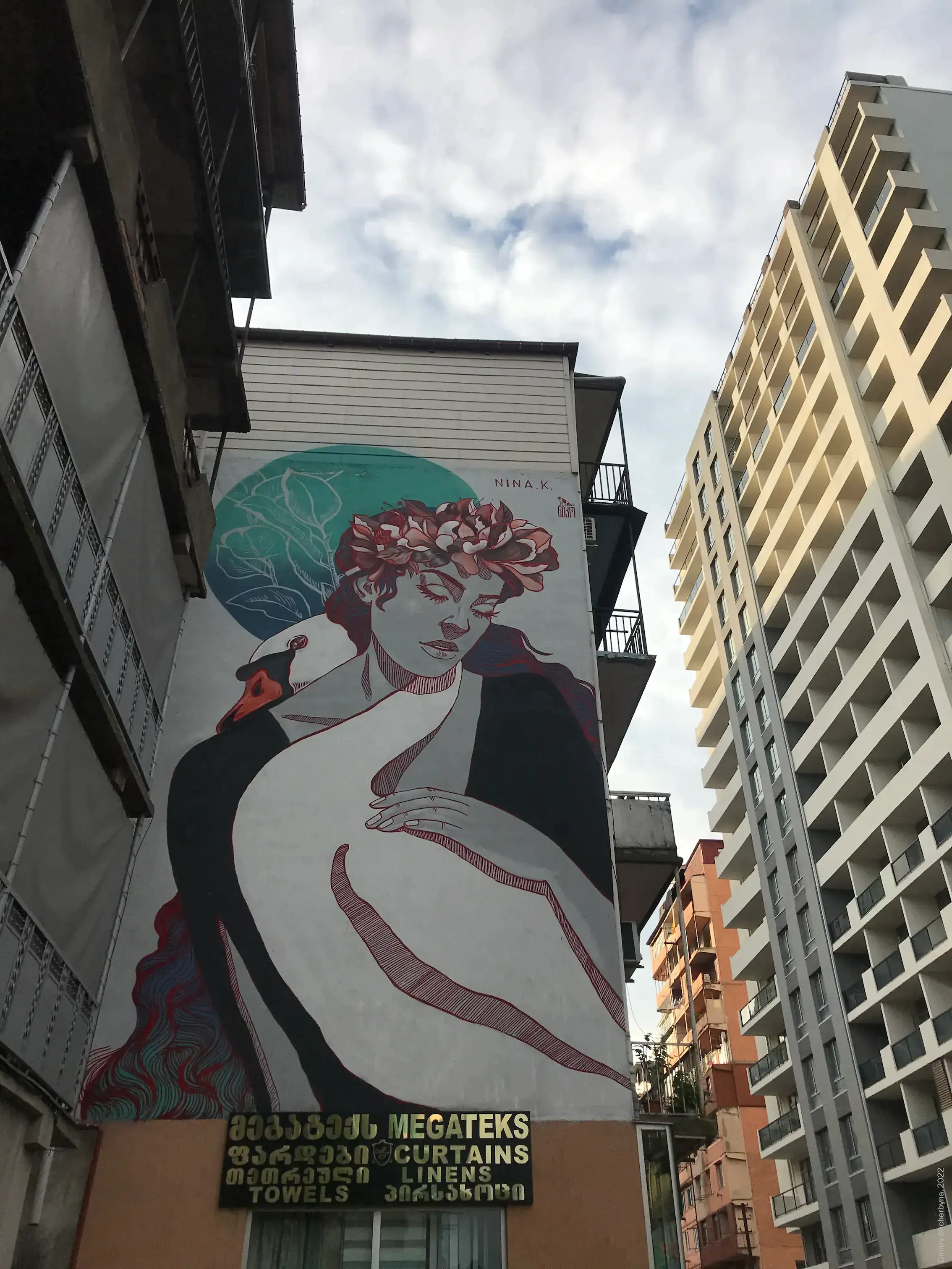 A mural of a woman with closed eyes and flowers in her dark red hair, gently embracing a white swan against a teal circular background. The style is inspired by art nouveau with fine red linework on pale skin.
