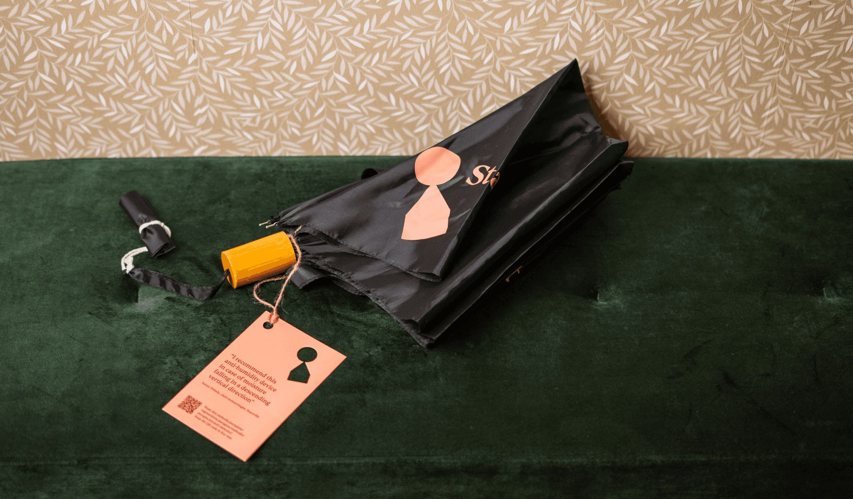 A black half-closed umbrella with a pink tag with a ouncheed out keyhole in the shape of the Staysville logosymbol and the text "I recommend this anti-humidity device in case of moisture falling in a descending vertical direction" and a QR code on it. The umbrella is laying on top of a green pouf next to a wallpapered wall with a leaf pattern in white. 