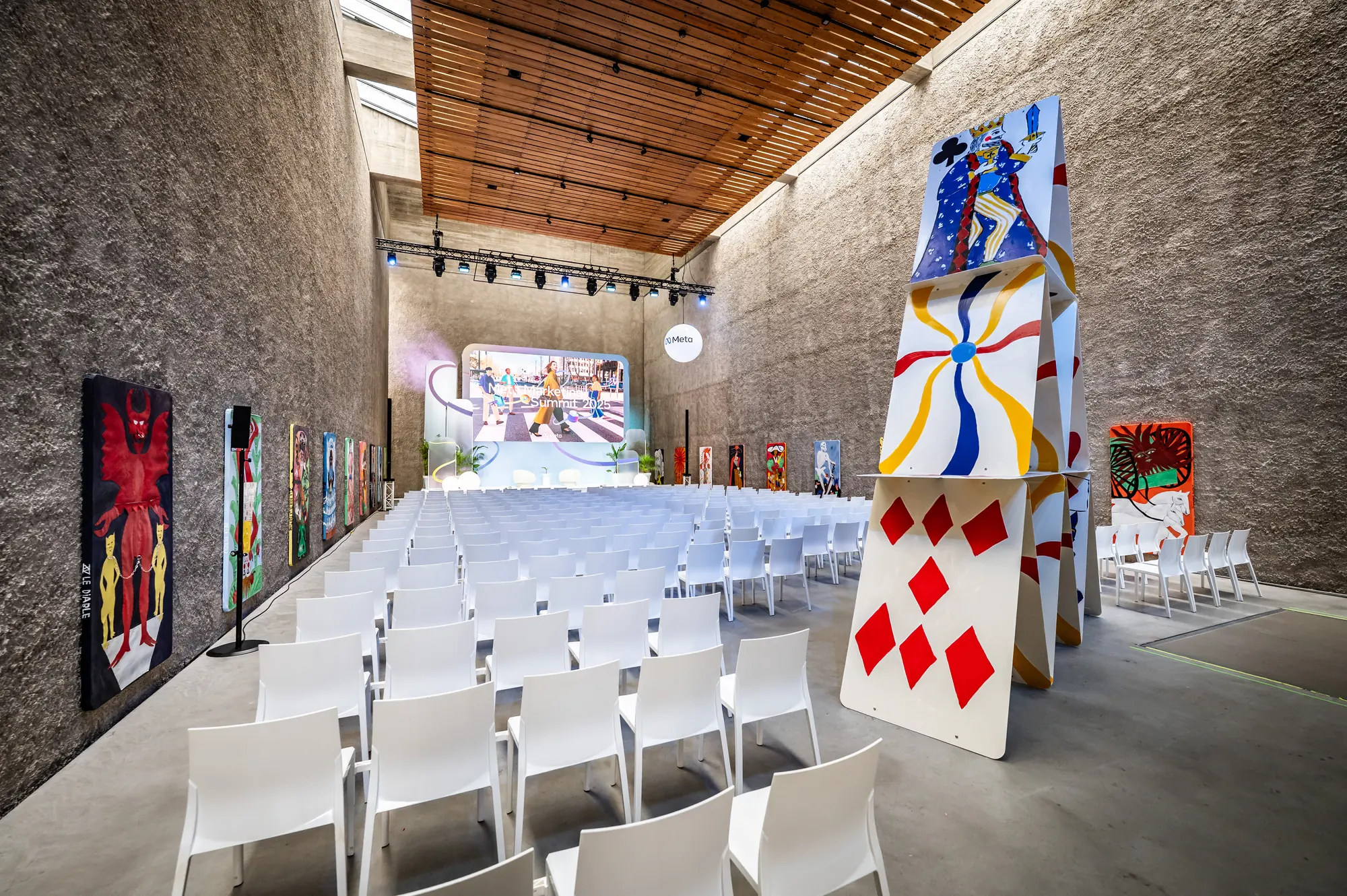 Conference hall with rows of white chairs and a wooden ceiling, surrounded by large-format art installations and patterned wall pieces at the Meta MMS event.