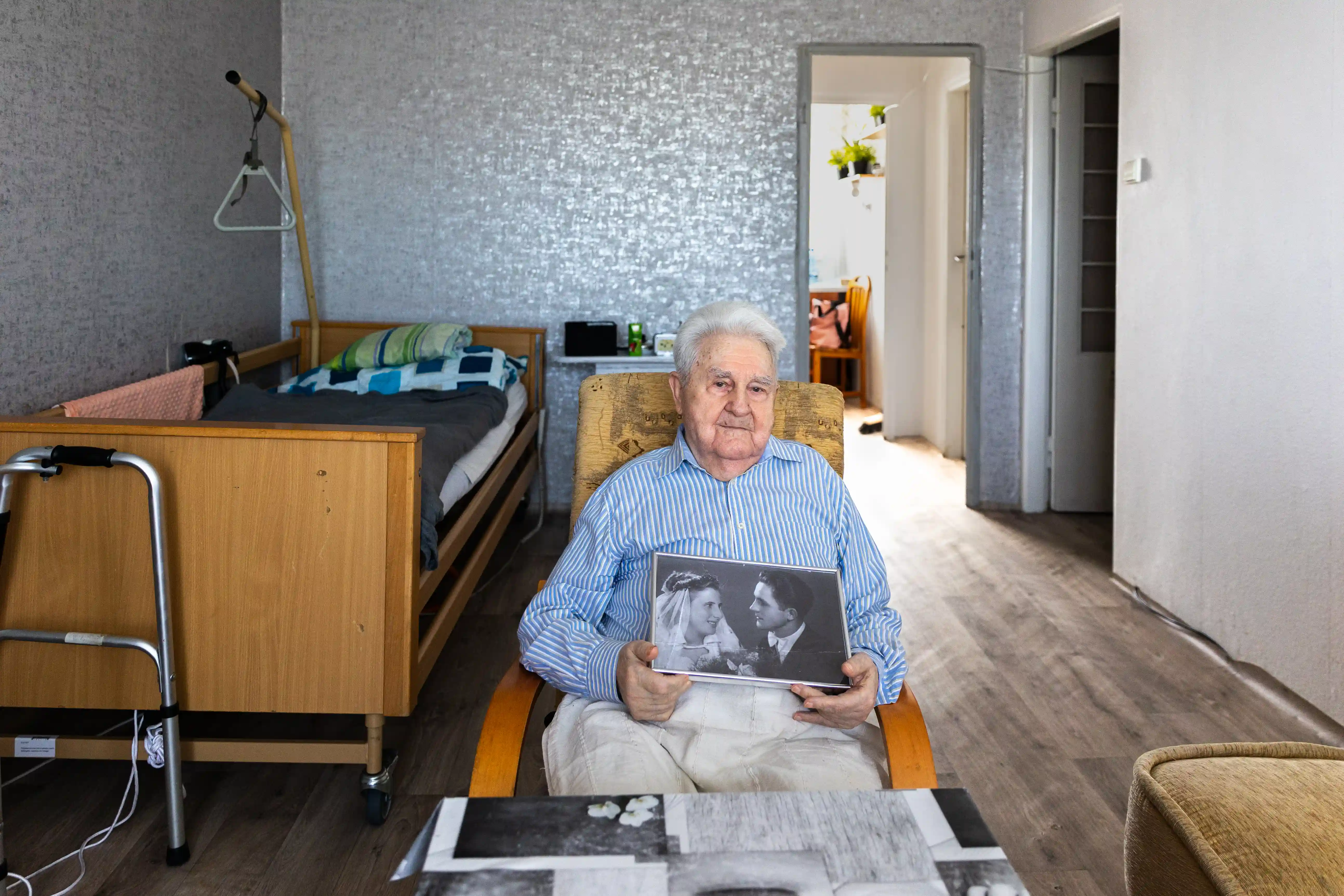 Starszy mężczyzna siedzący na fotelu trzyma w rękach czarno-białe zdjęcie małżeństwa. W tle sypialnia oraz chodzik.